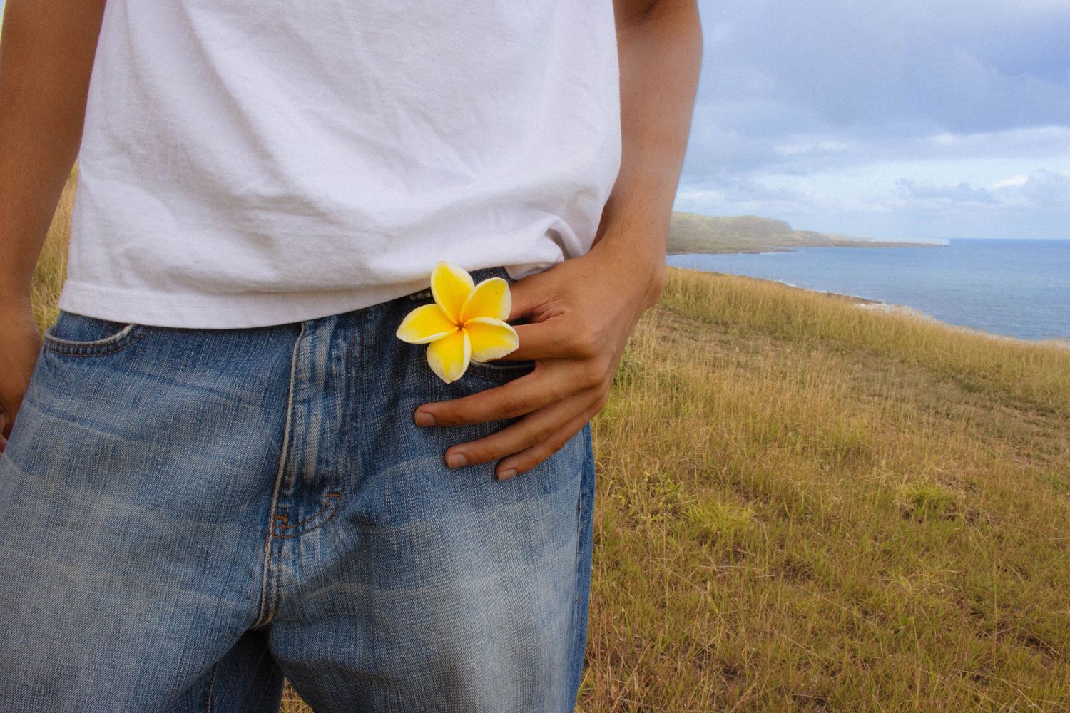 Close-up of a person in jeans holding a yellow plumeria flower with ocean in background.