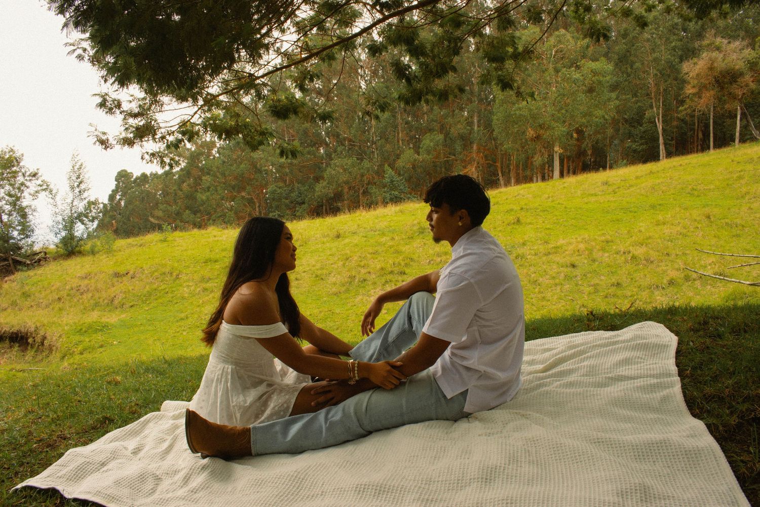 Young couple sitting face-to-face on a white blanket in a lush green park, sharing an intimate moment.