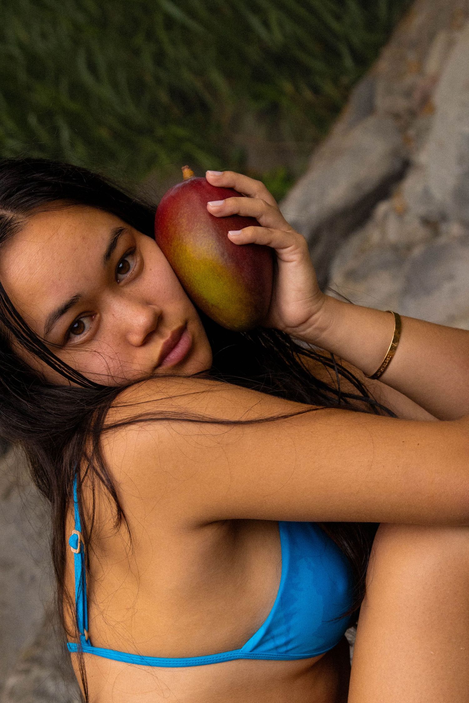 Young woman in blue bikini holding a mango against her cheek near rocks.