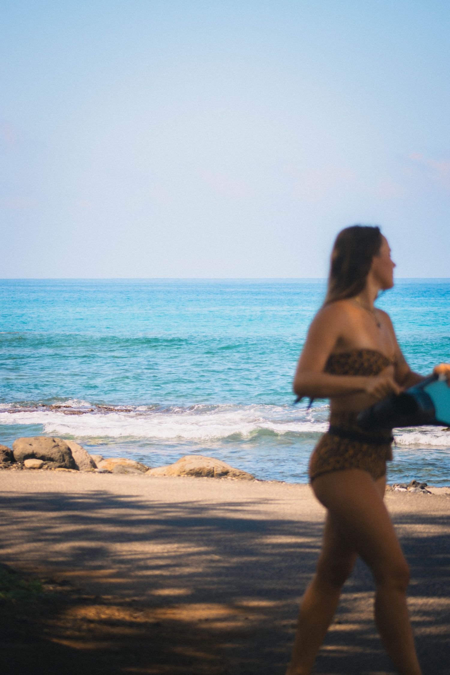 Blurred woman in leopard print bikini walking along a tropical beach with turquoise ocean water.