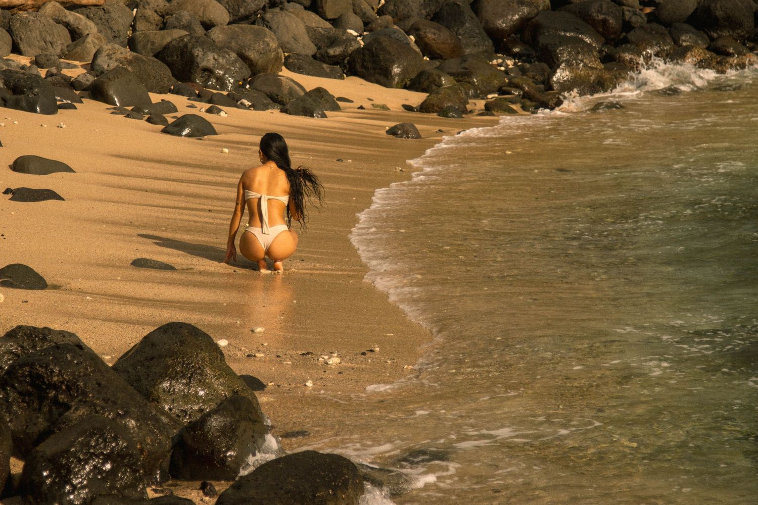 Woman in white bikini walking toward ocean waves on a rocky sandy beach.