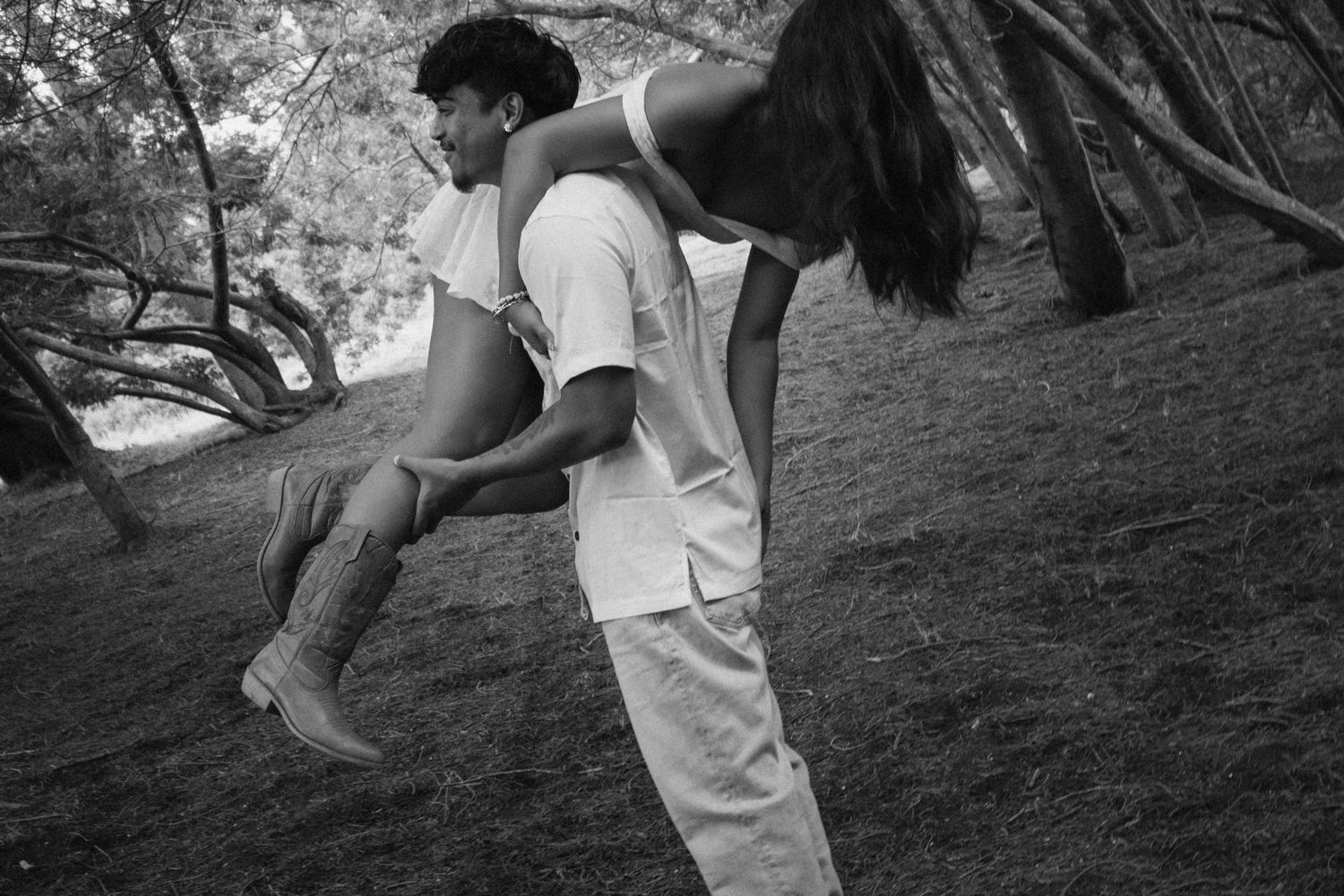 Black and white photo of a young man carrying a woman over his shoulder in a wooded outdoor setting.