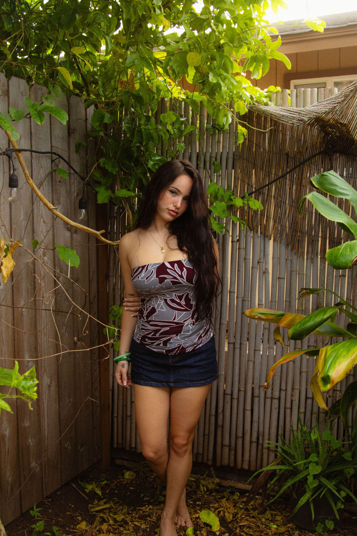 Young woman with long dark hair wearing a floral tube top and denim shorts standing by a bamboo fence.