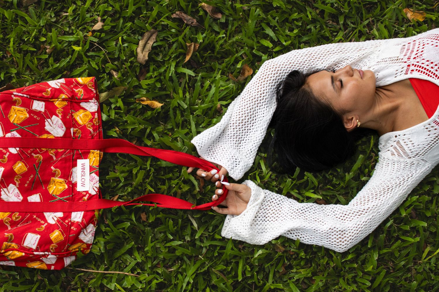 Woman lying on green grass holding a red floral Winslow tote bag overhead.