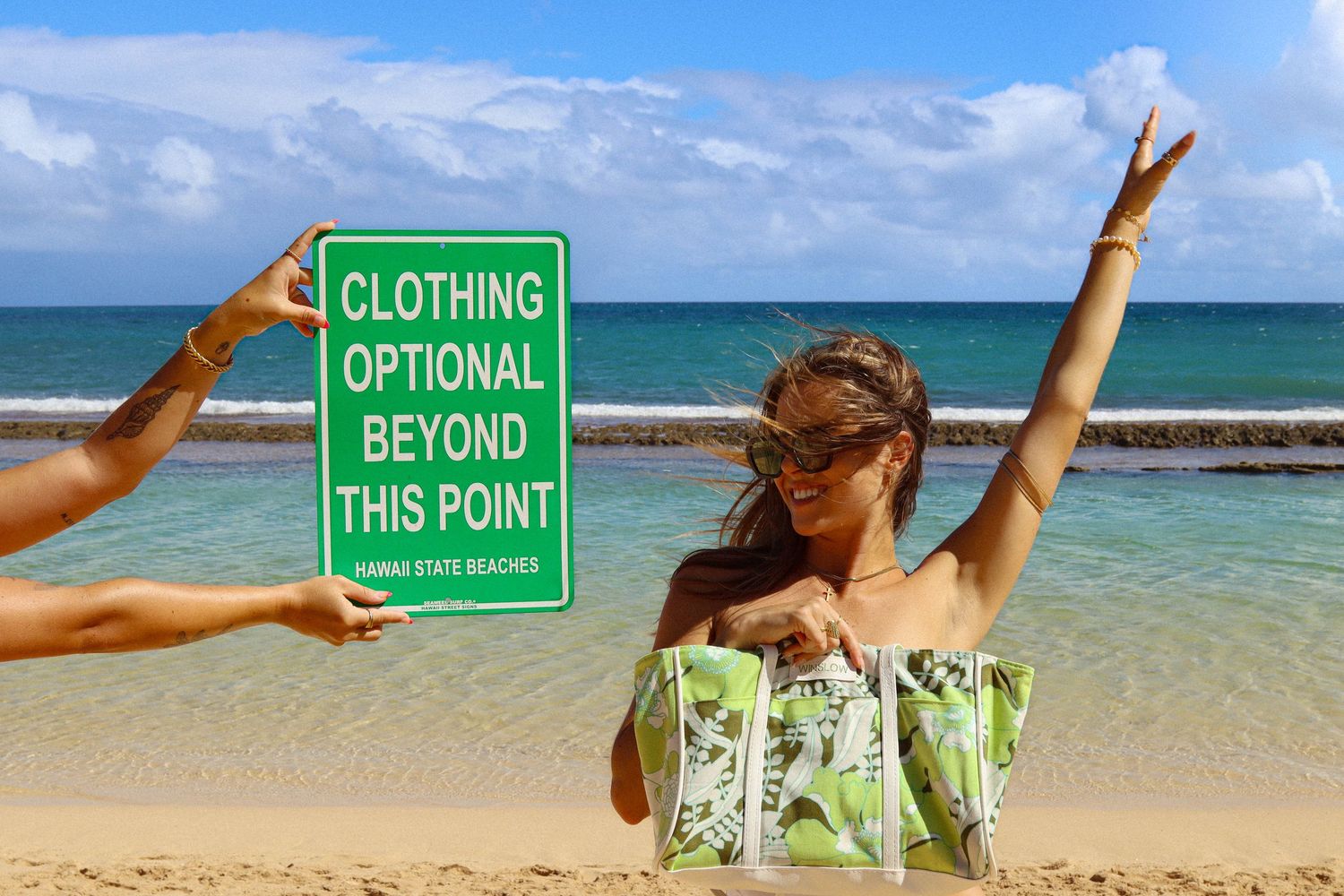 Woman at a beach holding a green clothing optional sign while another woman poses joyfully behind her.