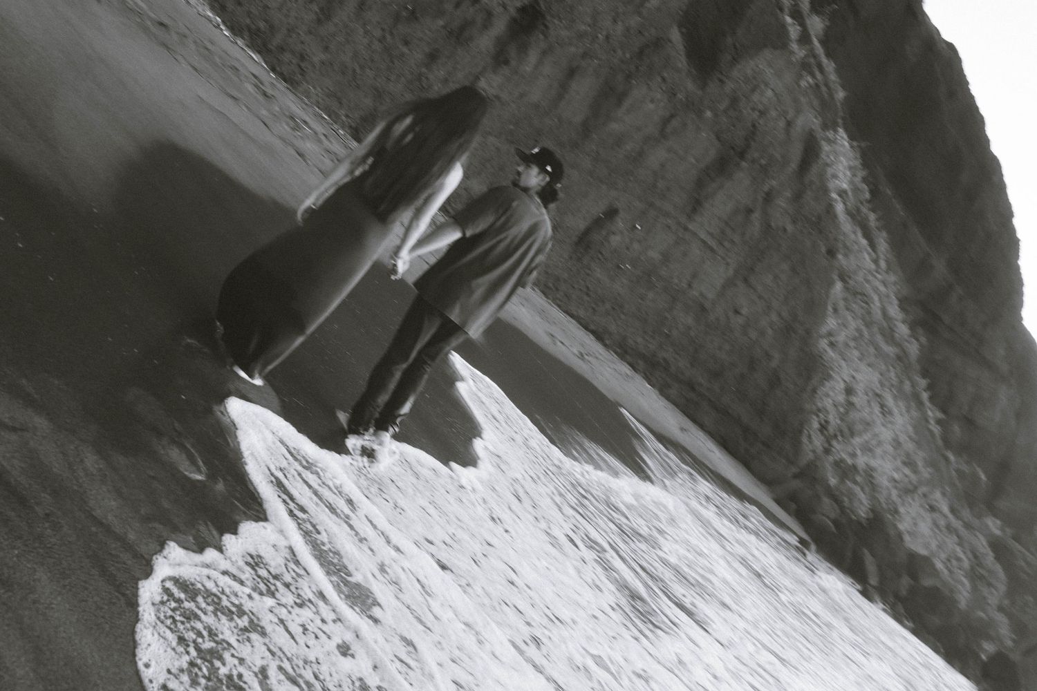Black and white photo of a person standing on a rocky beach with ocean waves.