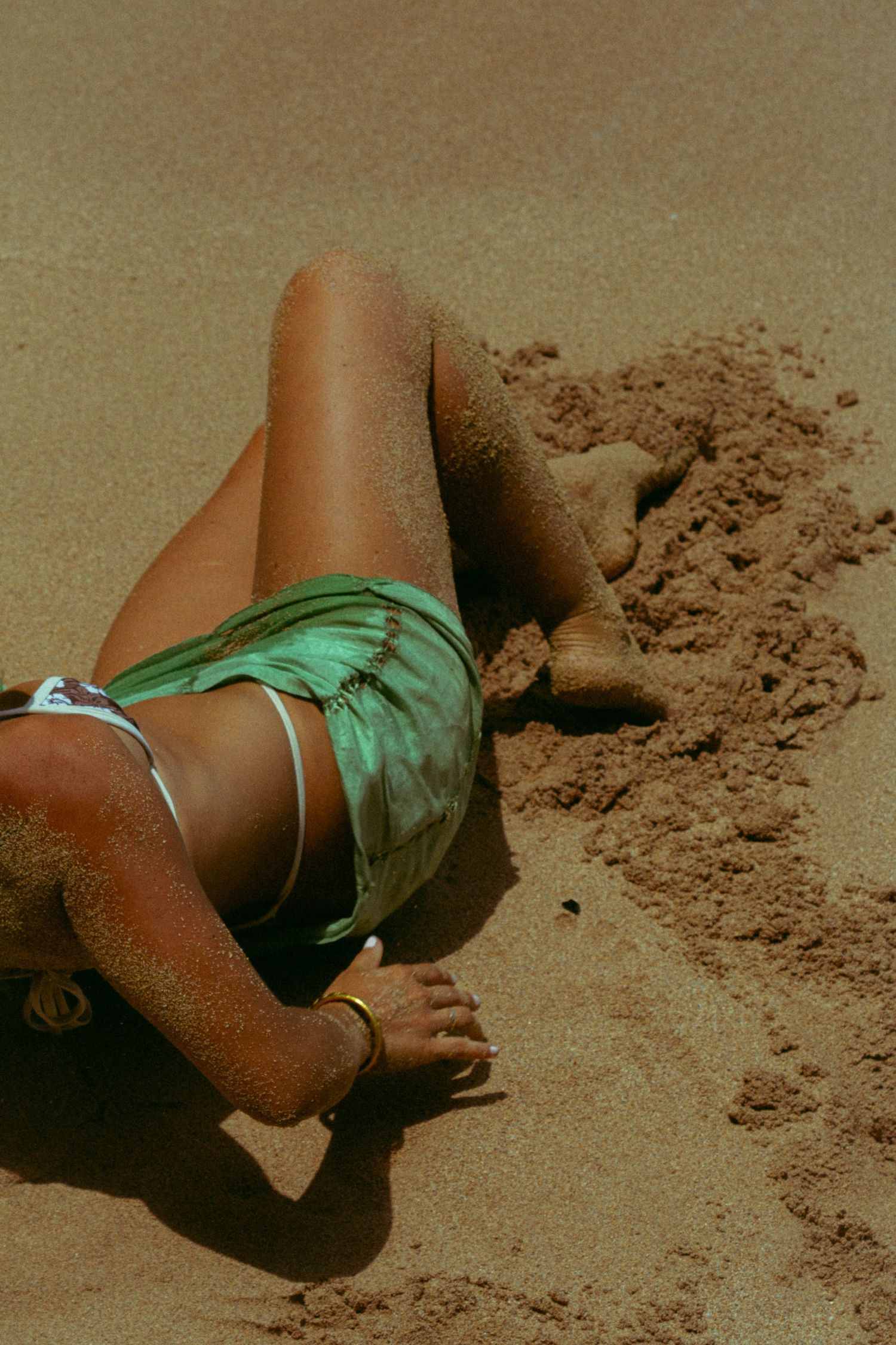 Close-up of tanned woman lying on sandy beach wearing green bikini bottoms and ankle bracelet.