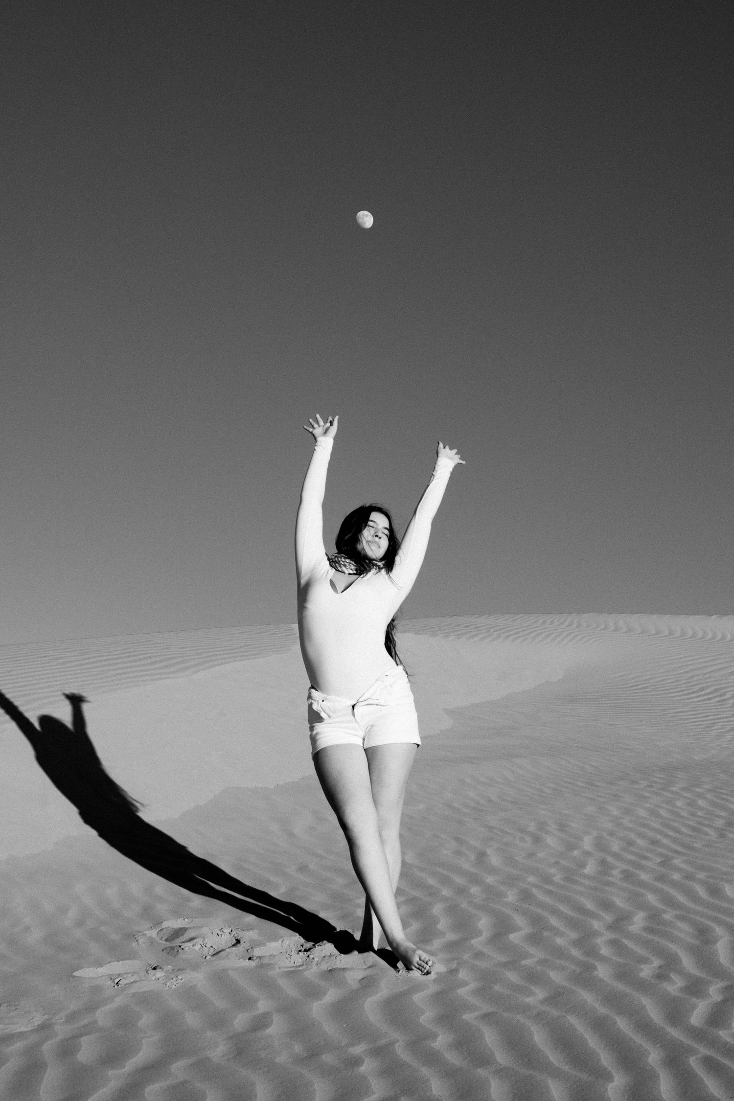 Black and white photo of a woman in shorts tossing a ball on desert sand dunes.