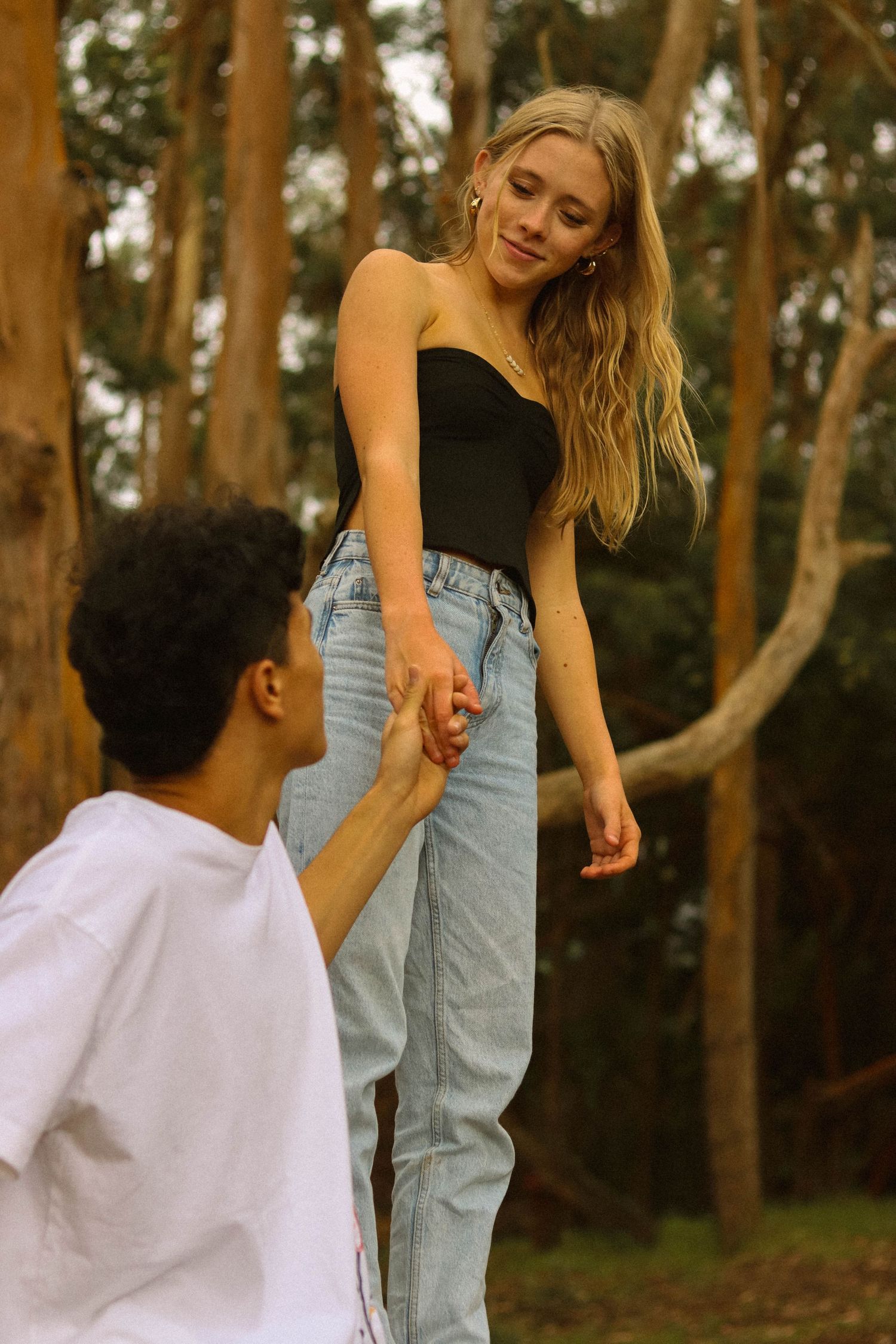 Blonde woman in black top and jeans holding hands with a man in a forest setting.