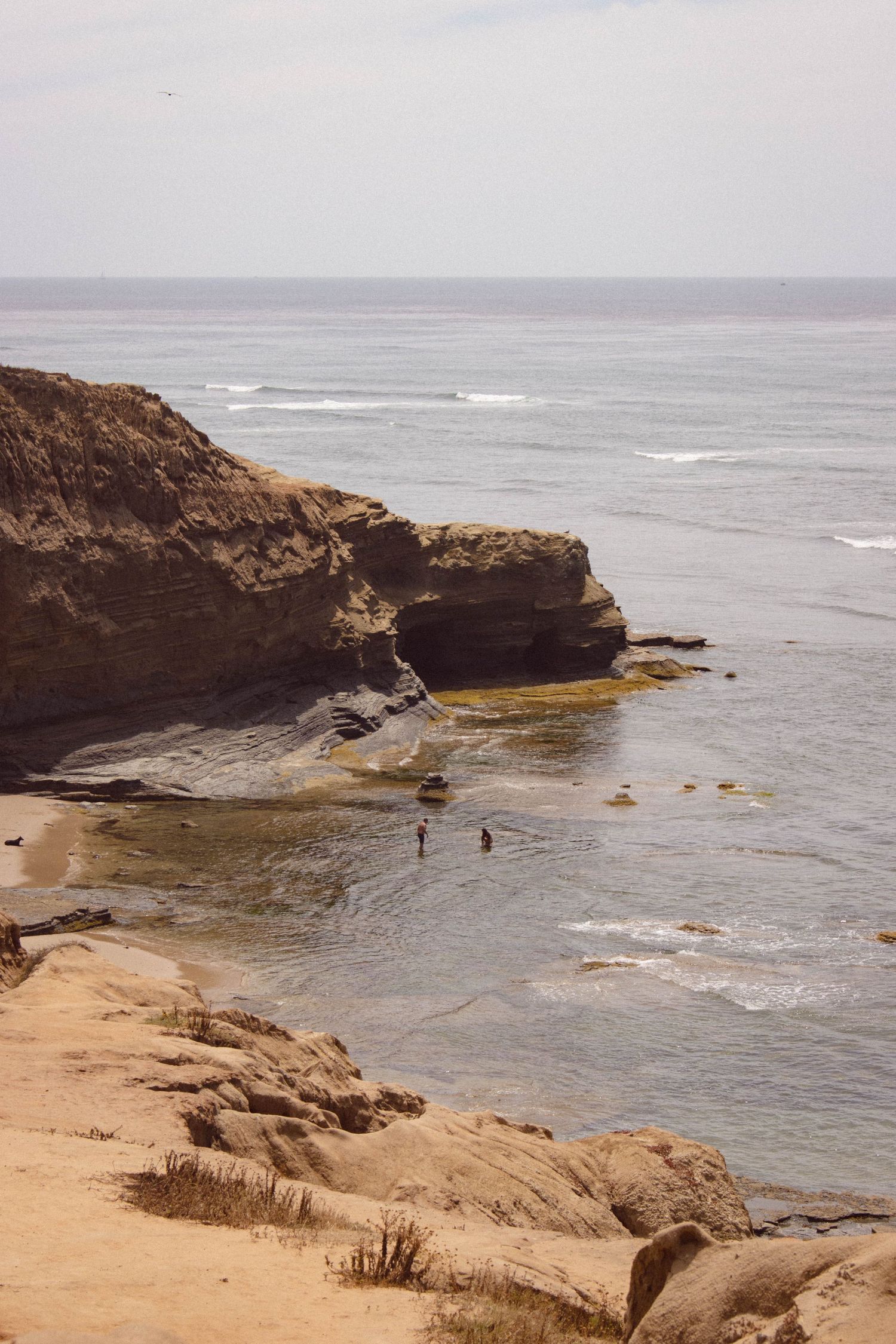 Rocky coastal cliffs with small cove beach and two people wading in calm ocean water.