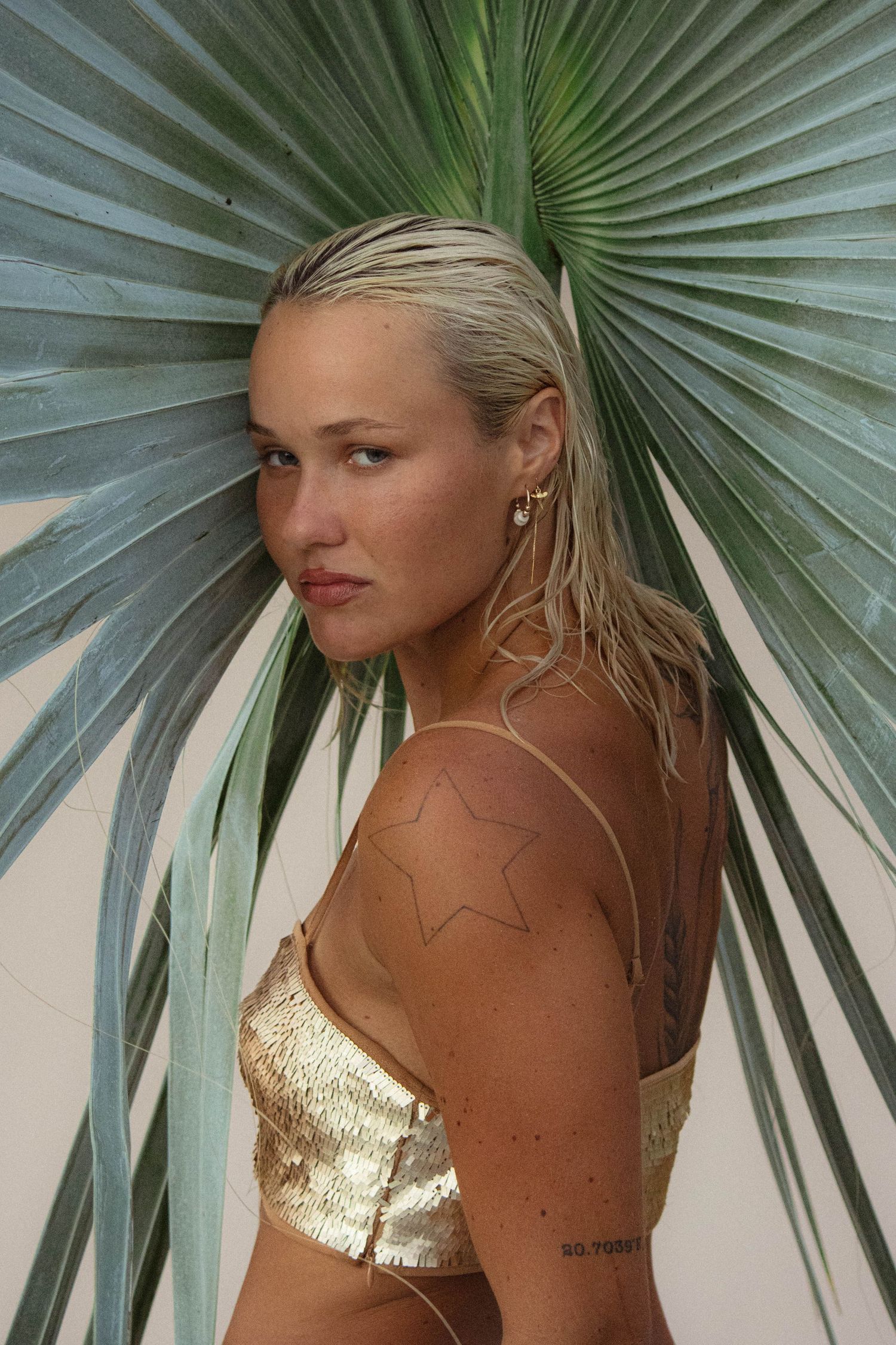 Blonde woman in gold metallic top posing against large green tropical palm leaf outdoors.