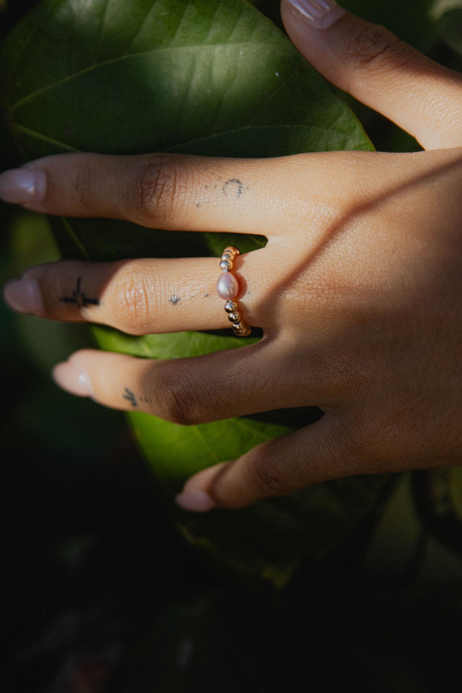 Hand adorned with a gold twisted pearl ring resting on lush green tropical leaves.