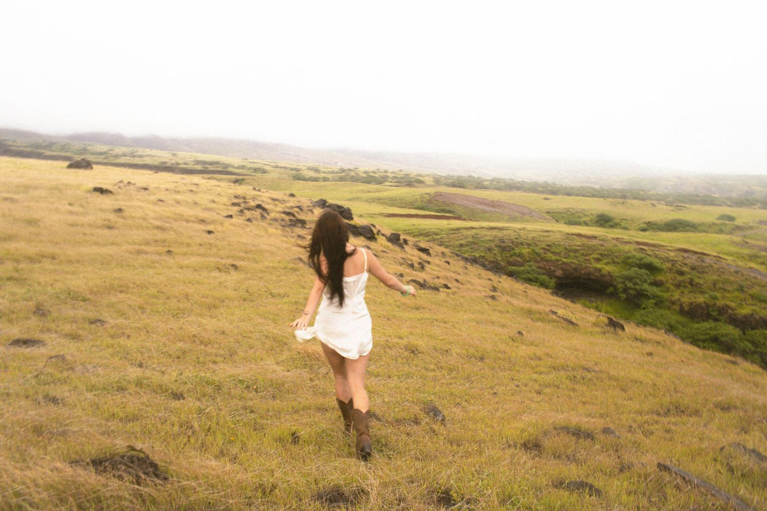Woman in white dress and boots walking through green meadow with scenic foggy landscape in background.