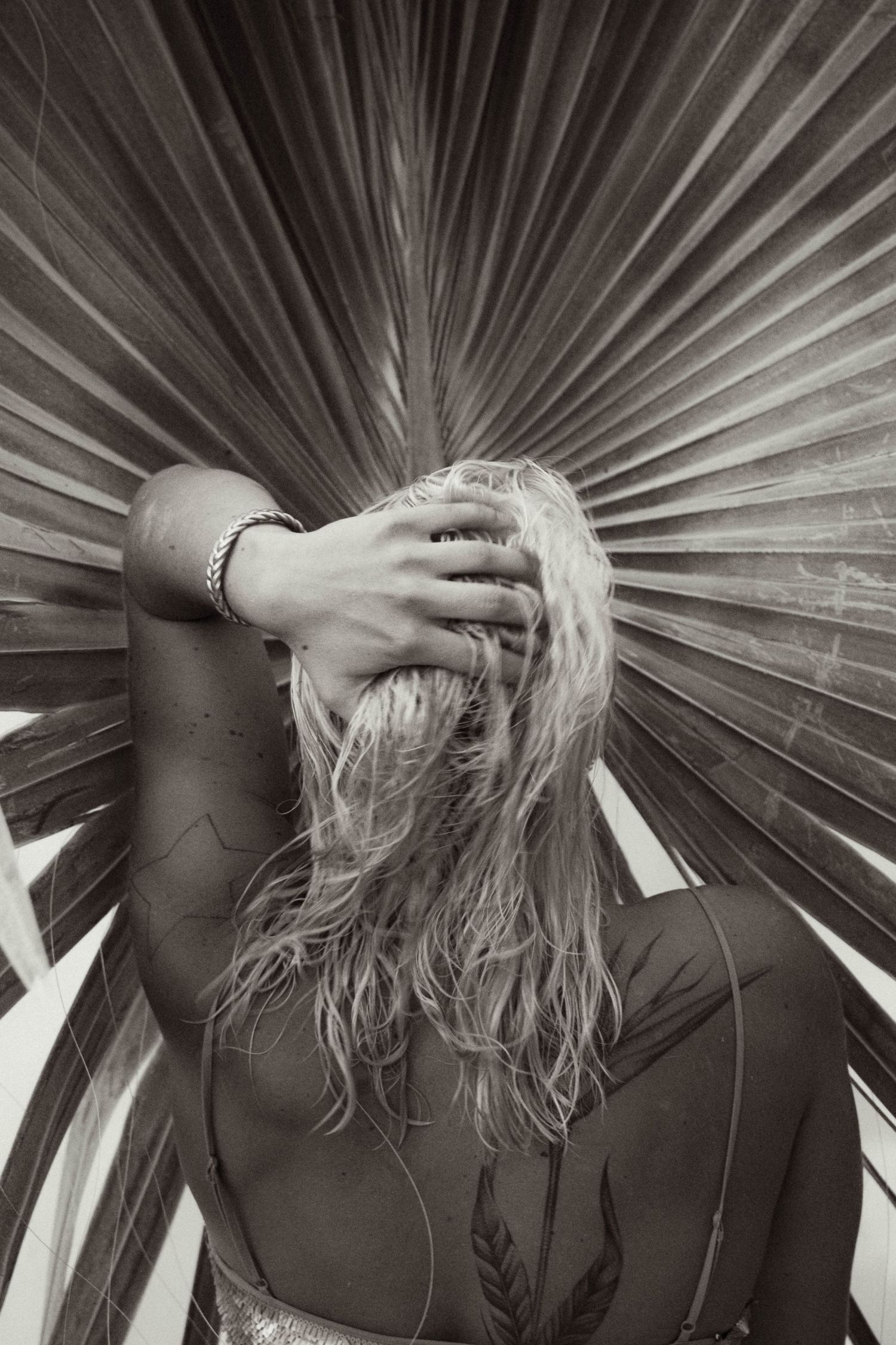 Black and white photo of blonde woman with back tattoo posing in front of large tropical palm leaf.