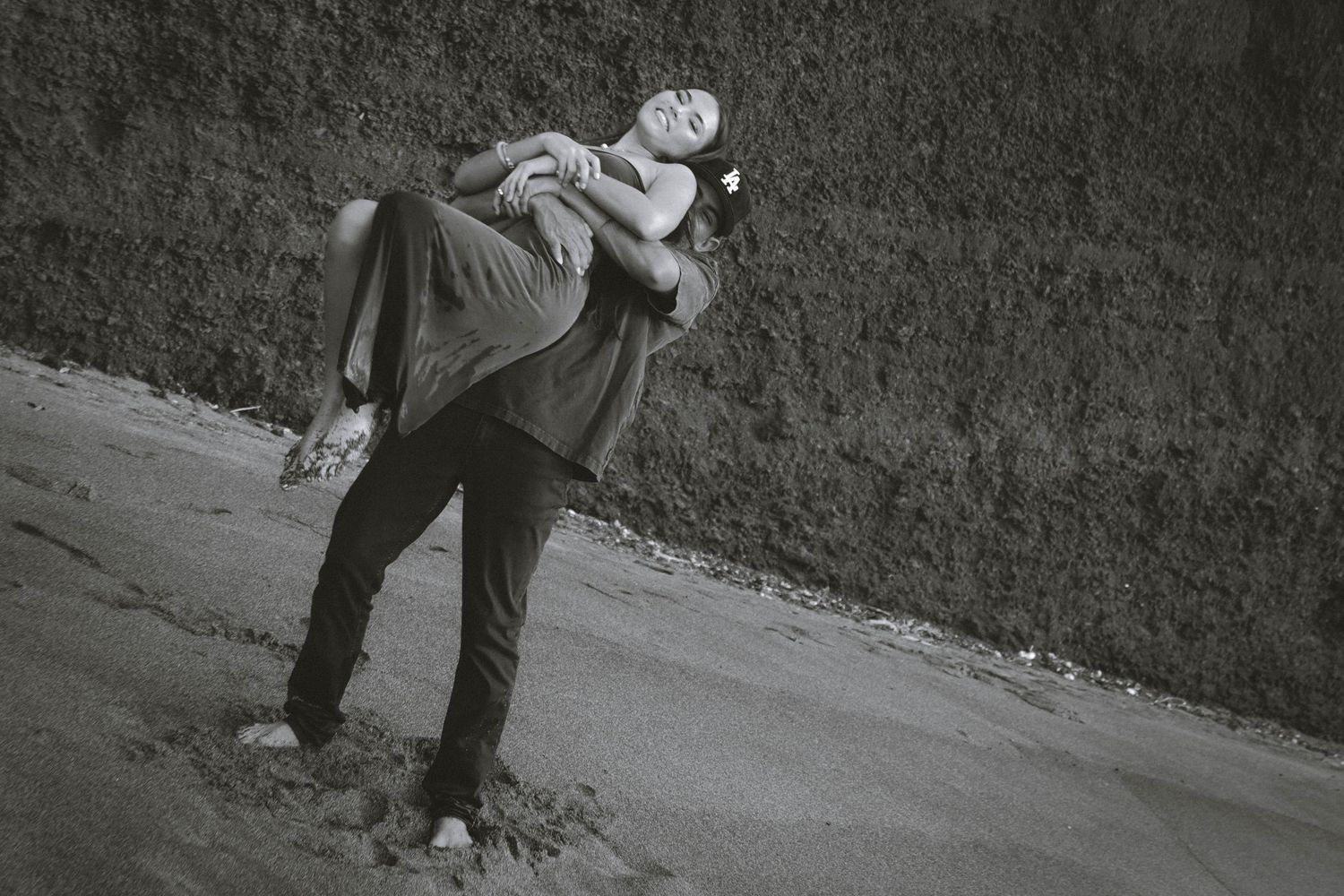 Black and white photo of a man lifting a woman outdoors near a textured wall.