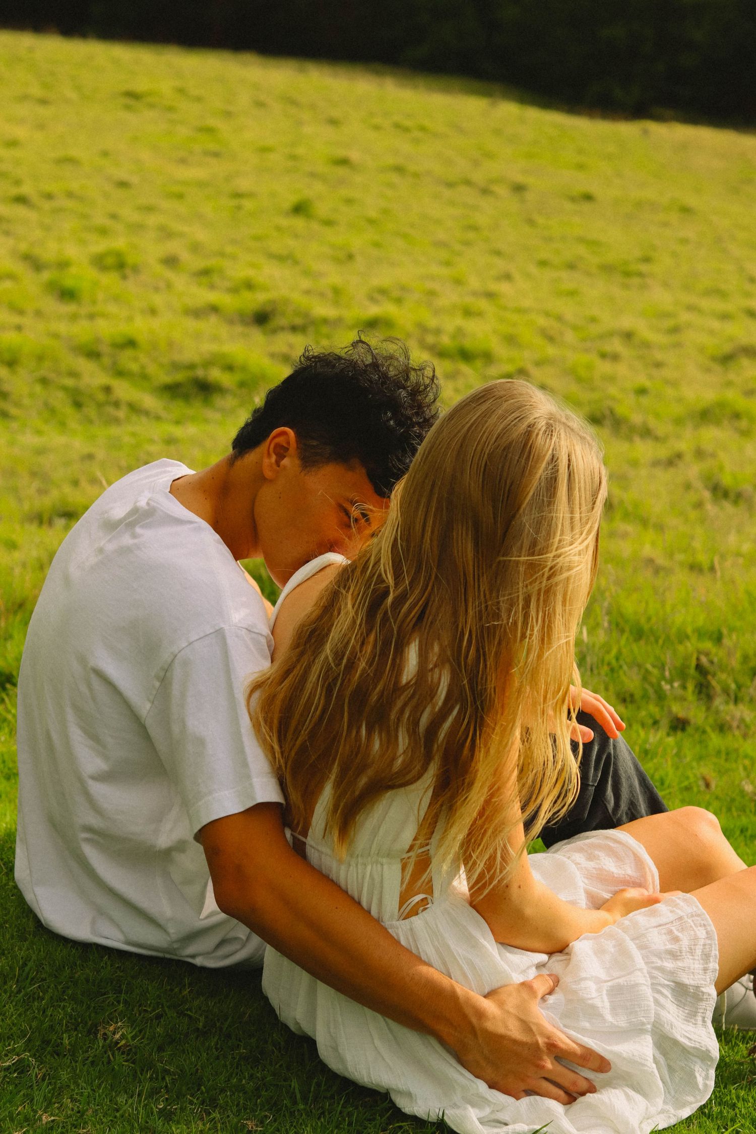 Young interracial couple sitting closely together on green grass hillside.