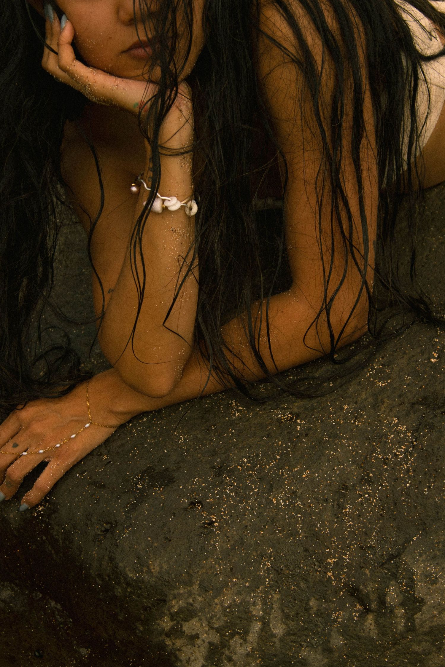 Woman with long wet dark hair lying on dark sand wearing silver jewelry.