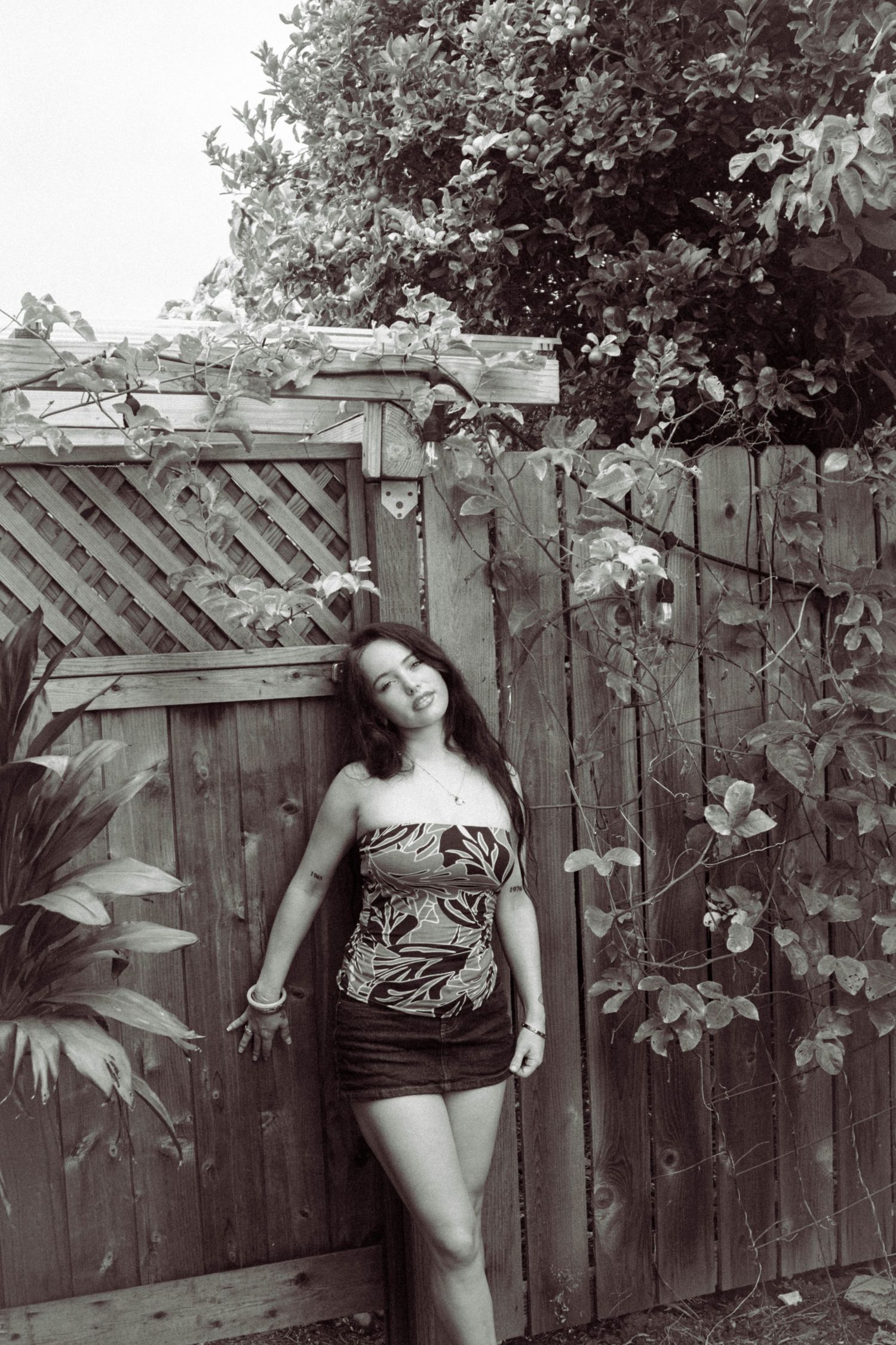 Black and white photo of a young woman in a floral tube top leaning against a wooden fence with vines.