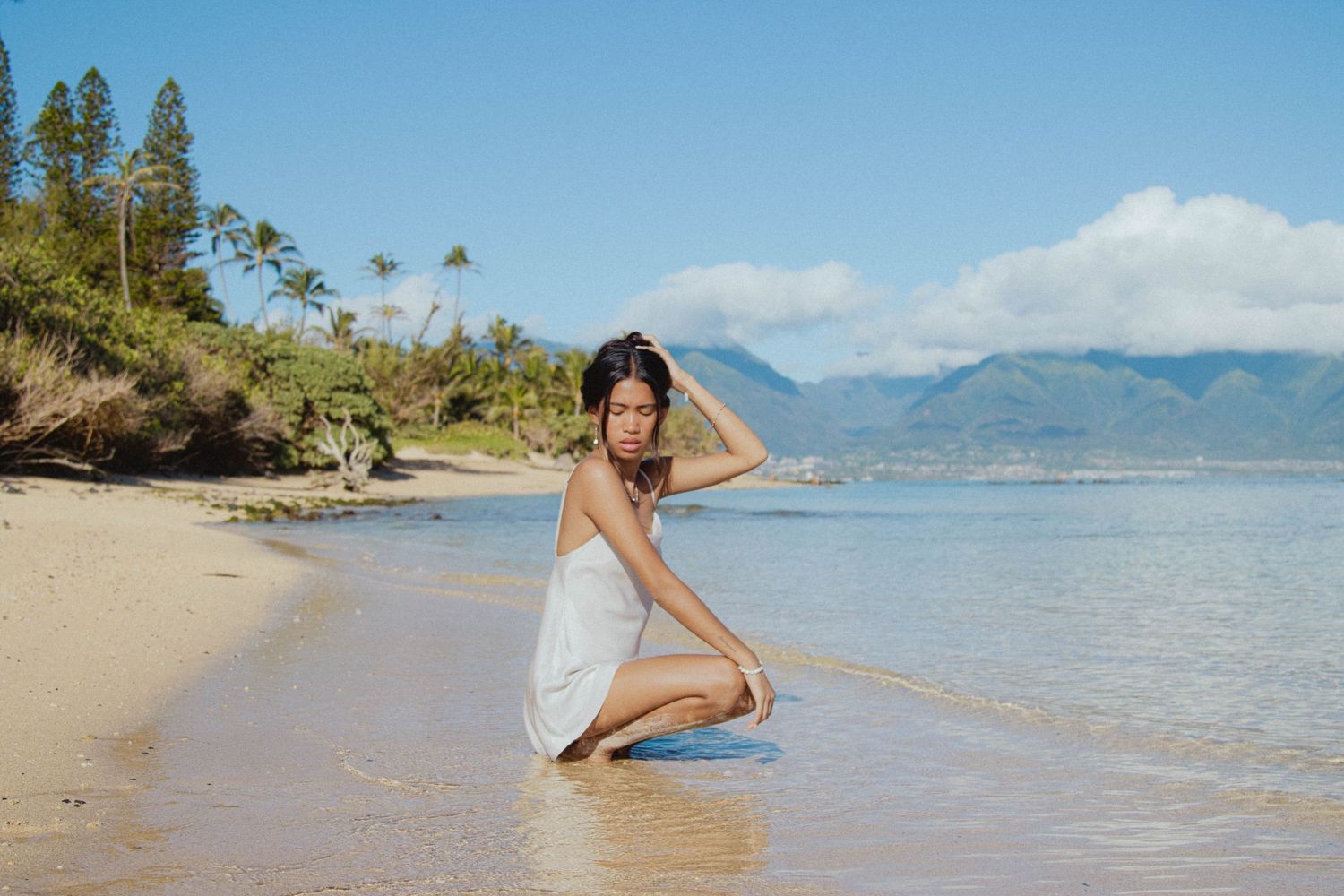 Young woman in white dress kneeling on a tropical beach with mountains in the background.