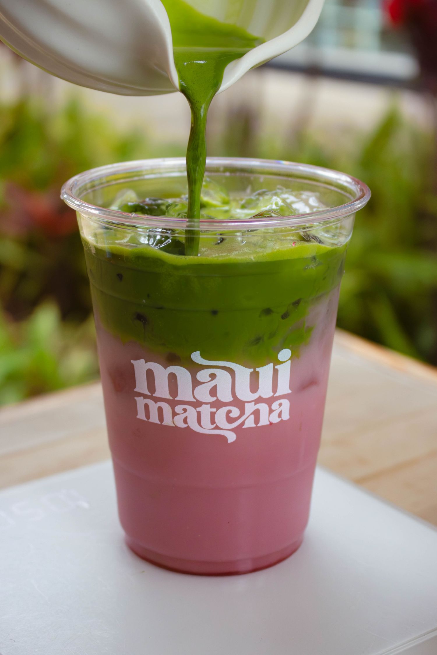 Green matcha being poured over pink milk and ice in a Maui Matcha branded clear plastic cup.