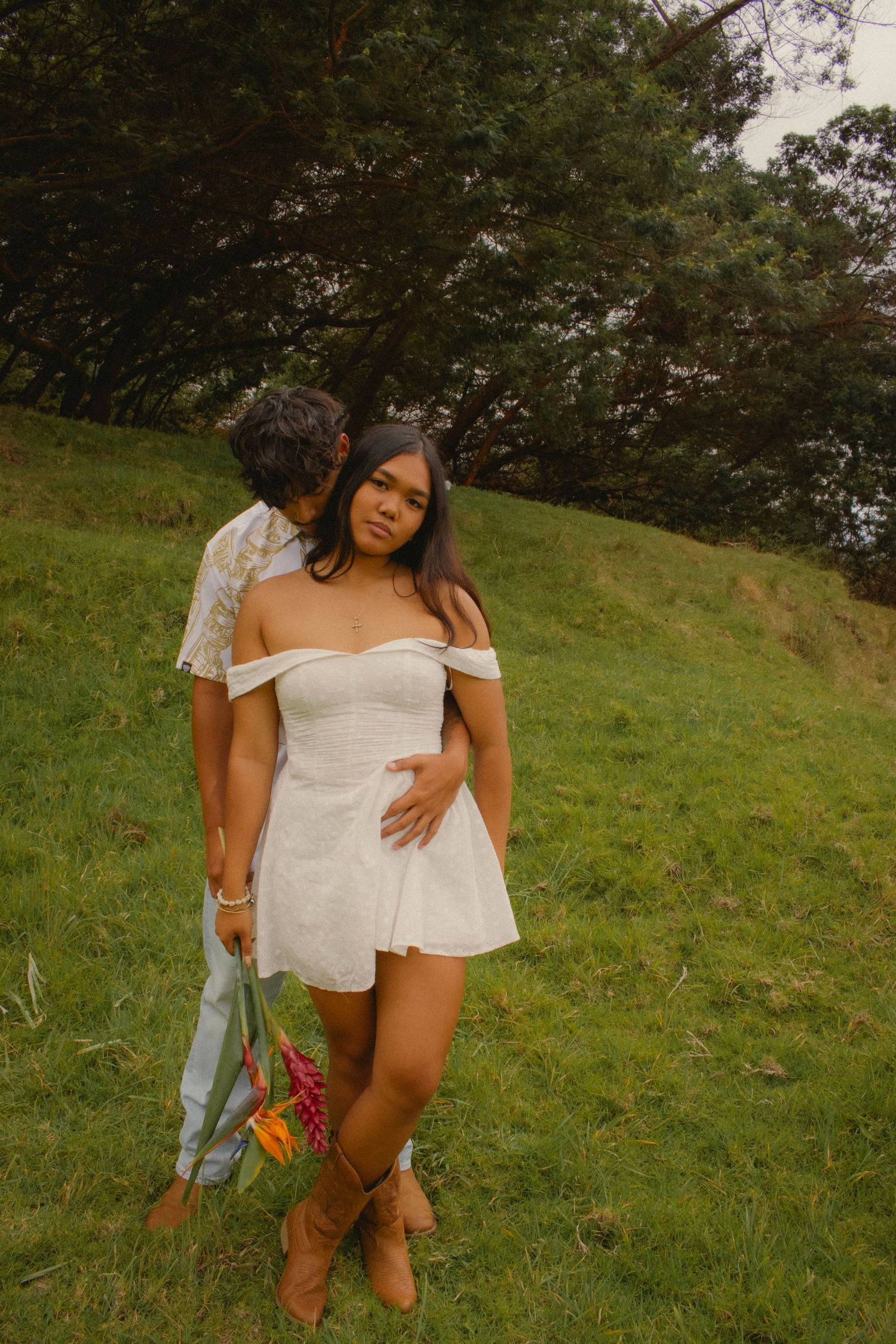 Young woman in a white off-shoulder mini dress and brown boots standing on a grassy hill with a man.