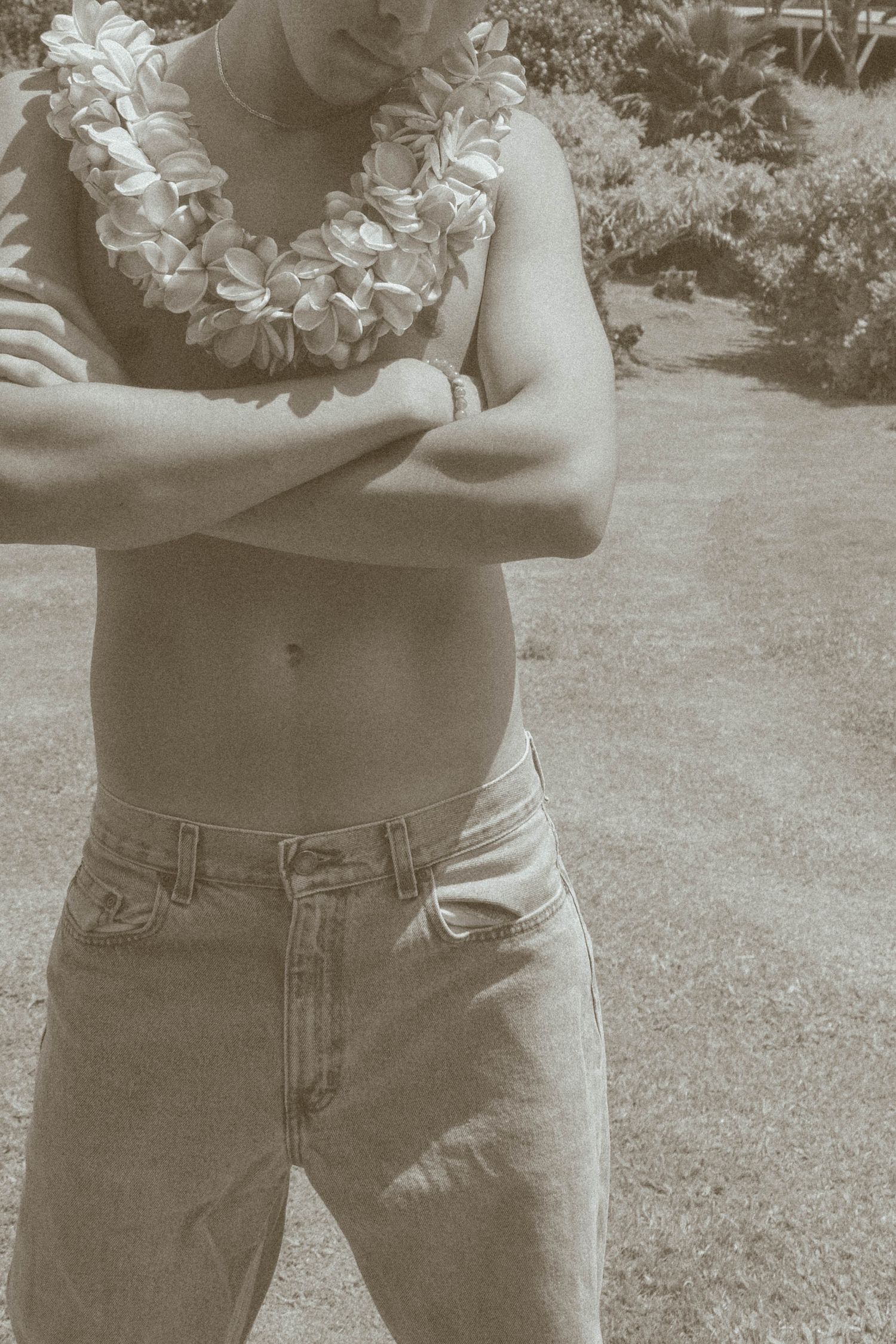 Sepia-toned photo of a shirtless man wearing a floral lei with arms crossed outdoors.