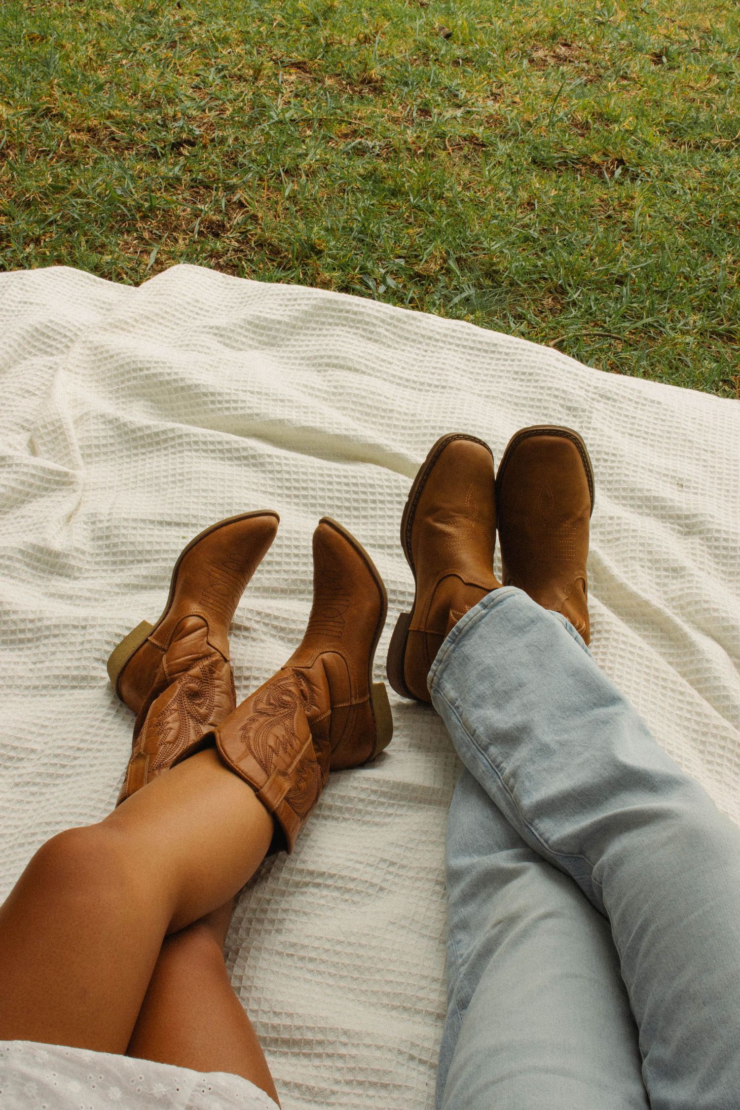 Two people wearing matching brown cowboy boots relaxing on a white blanket over green grass.
