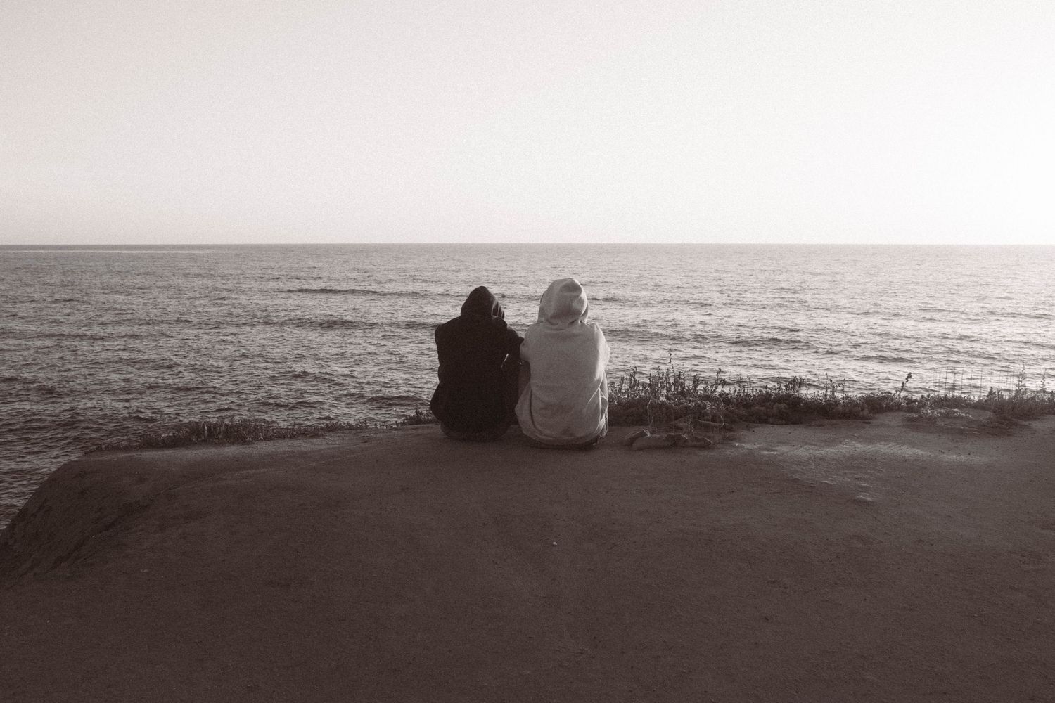 Two people sit side by side on a rocky cliff overlooking a calm ocean horizon in black and white.