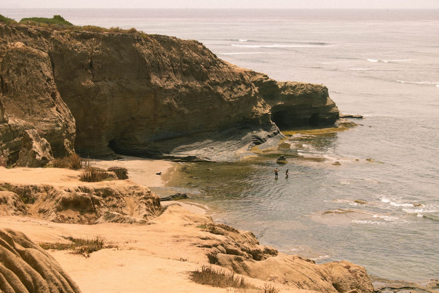 Rocky coastal cliffs frame a secluded sandy cove with two people wading in the ocean below.