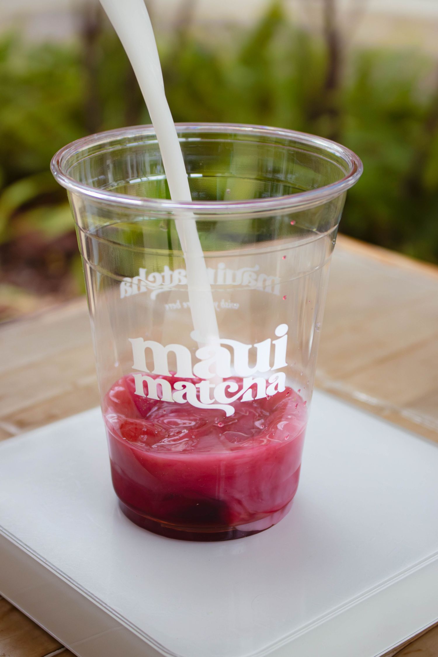 Red berry drink being poured with milk into a Maui Matcha branded clear plastic cup on a white surface.