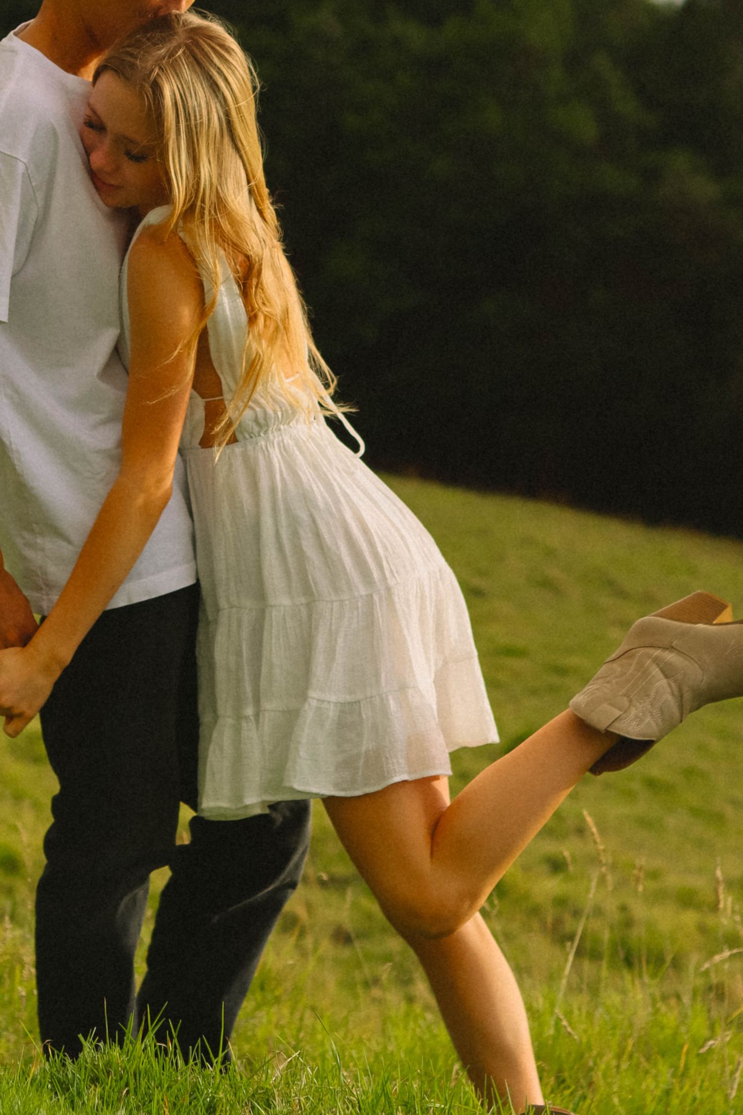 Couple embracing in a sunlit green meadow, woman in white dress kicking up her foot.