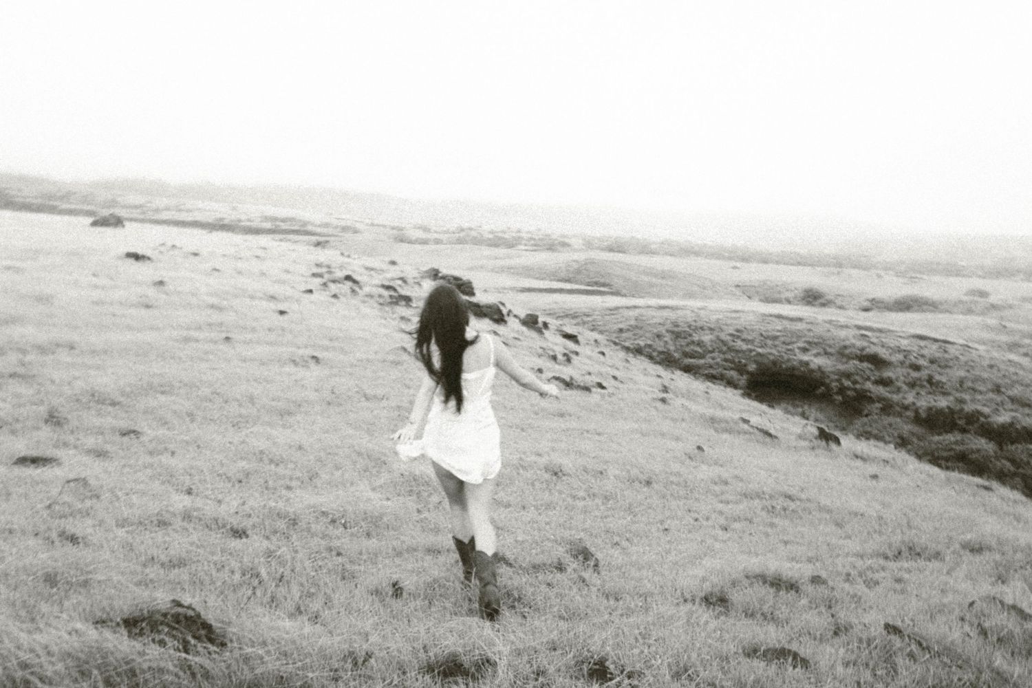 Black and white photo of woman in white dress and boots walking across grassy hilltop landscape.