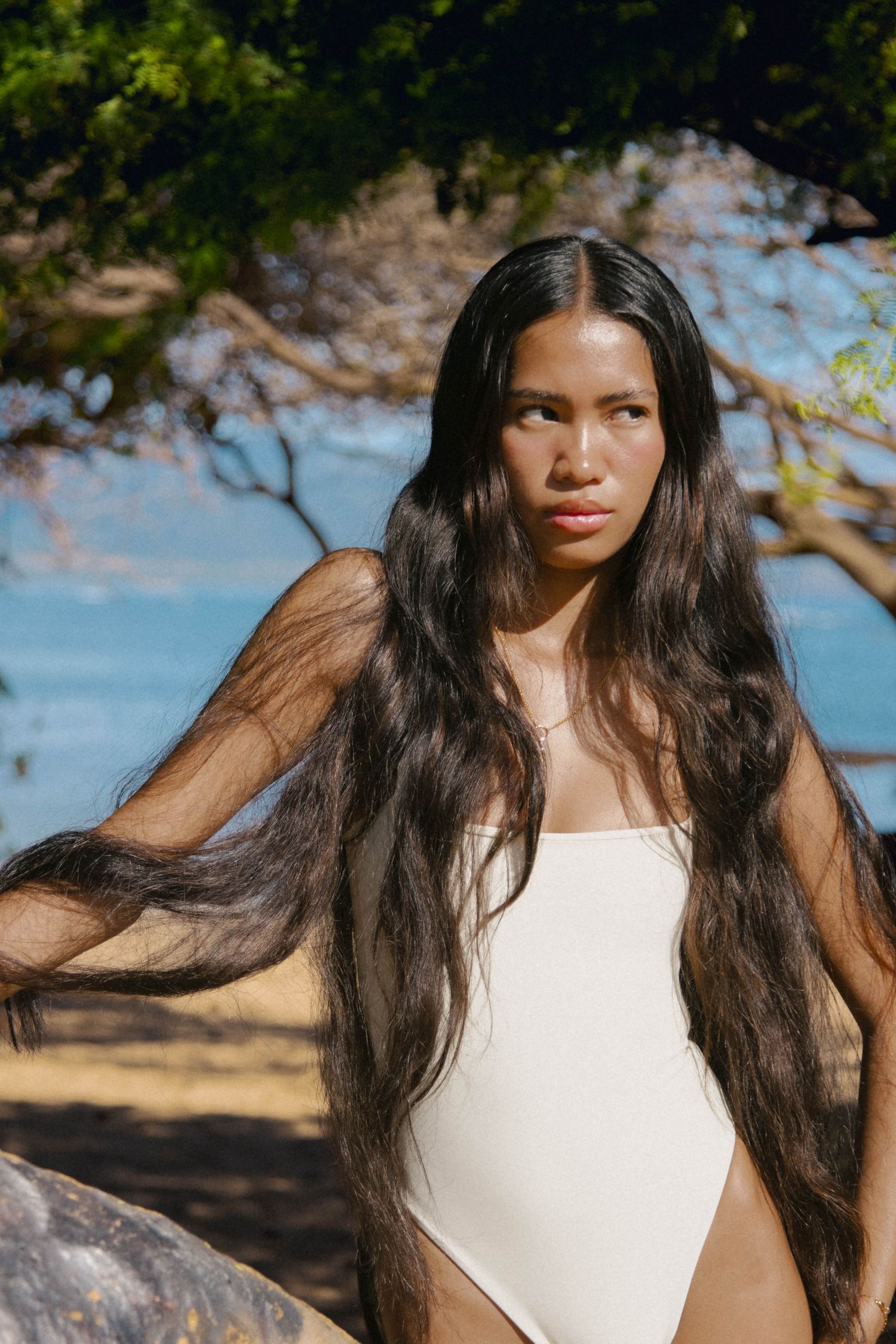 Woman with long flowing dark hair in white one-piece swimsuit posing near ocean shoreline trees.