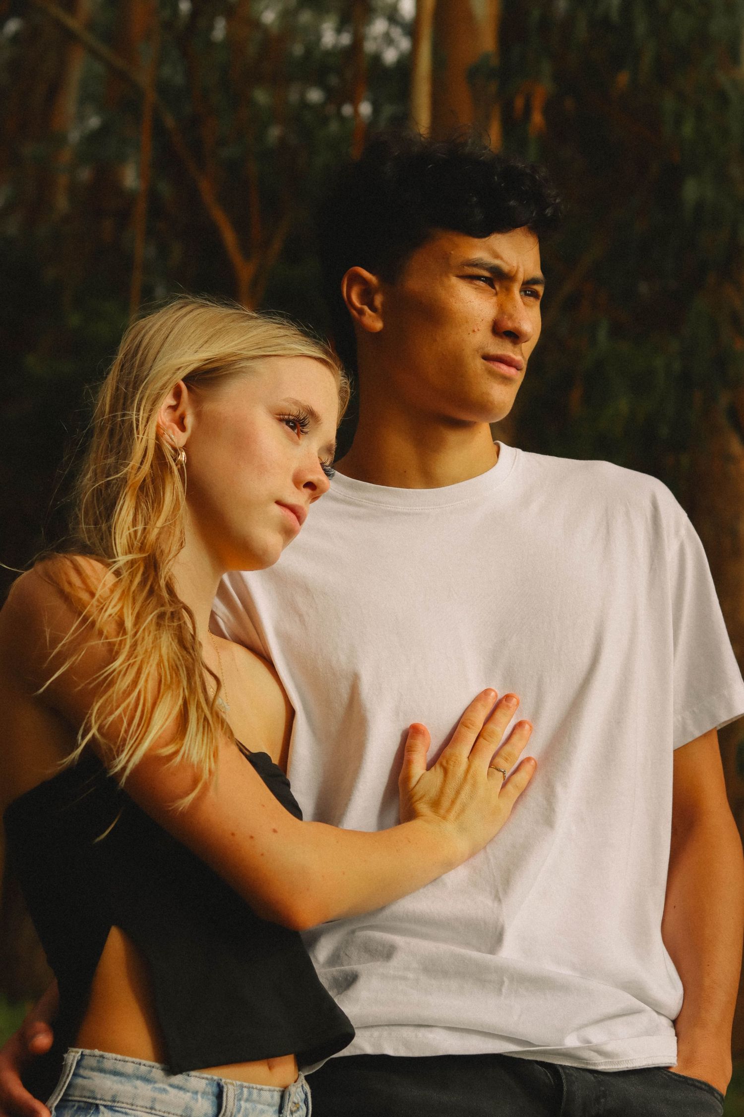 Young couple embracing outdoors, blonde woman leaning on mans white t-shirt in golden light.