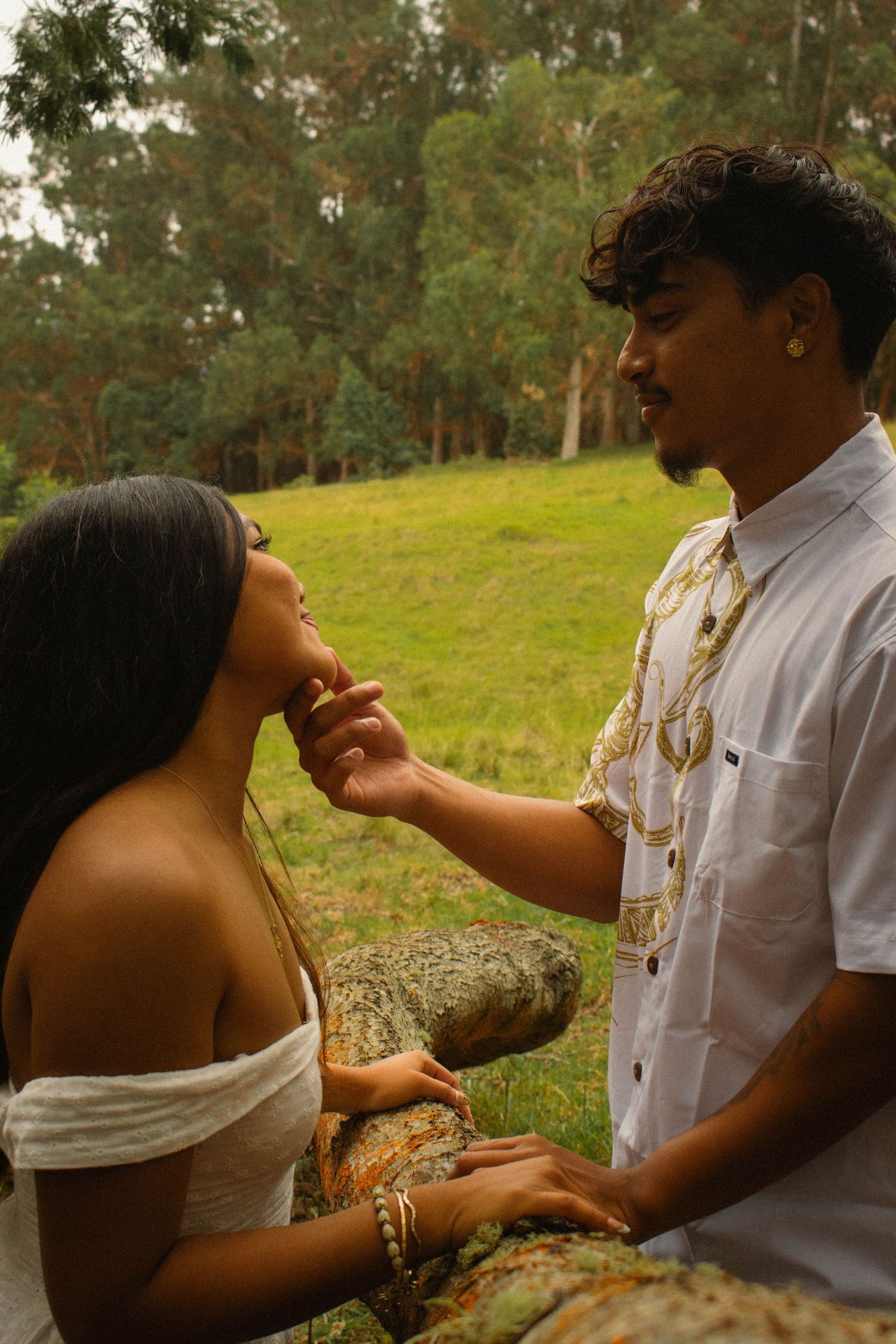 Young man tenderly touching a woman's face while she sits on a rock in a green forested park.