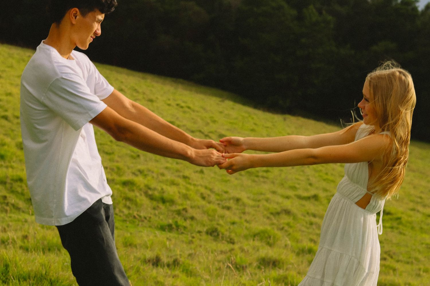 Young couple holding hands and spinning in a sunny green meadow wearing white outfits.