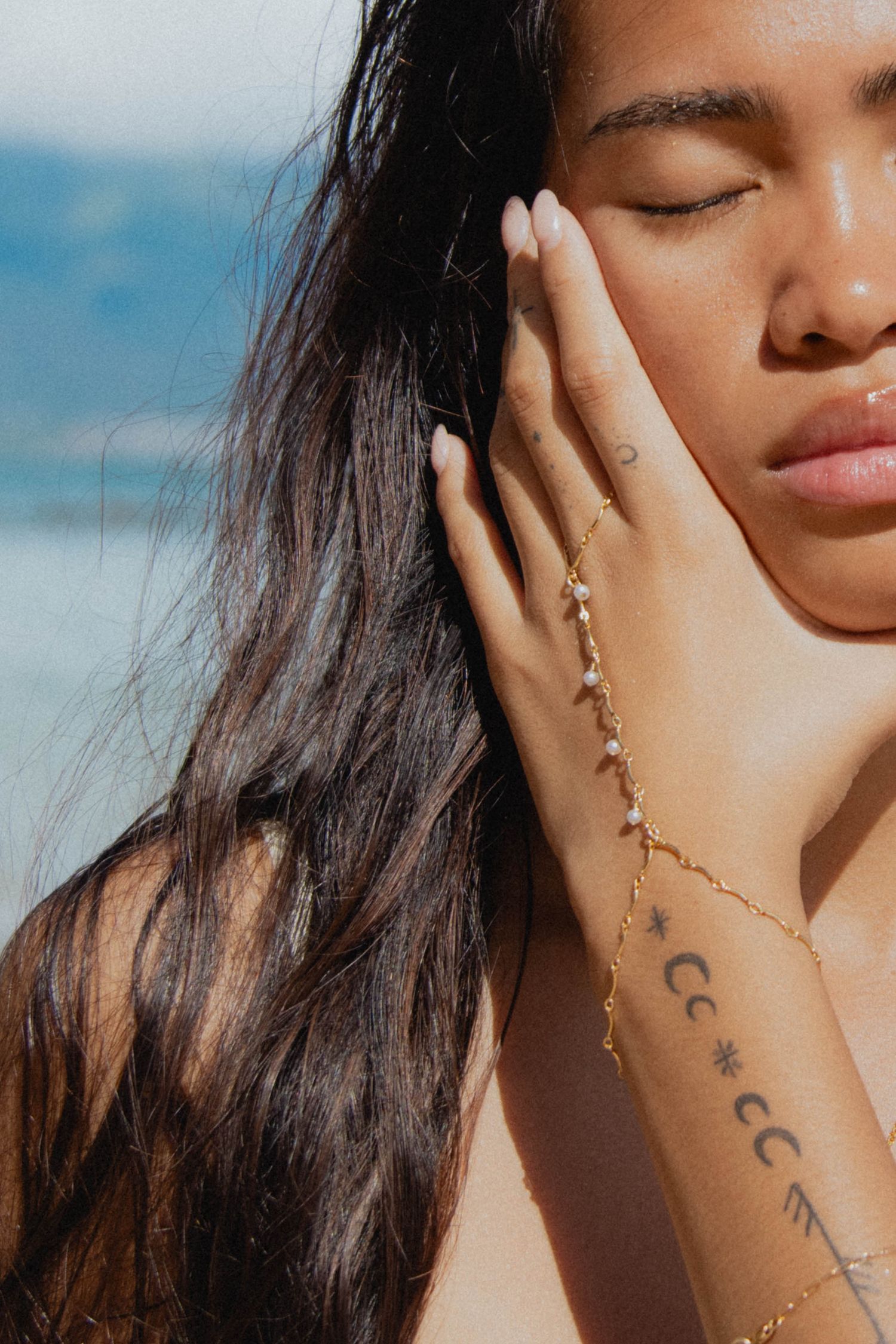 Young woman with closed eyes and dark hair touching her face, wearing a gold chain hand bracelet with tattoos visible.