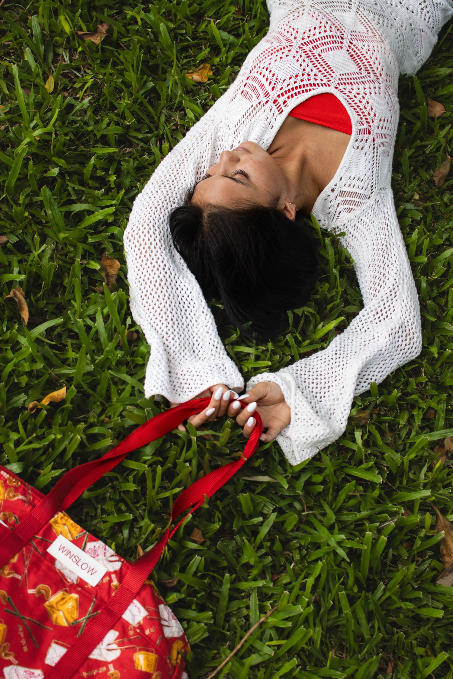Woman in white crochet dress lying on grass holding red Winslow printed tote bag.