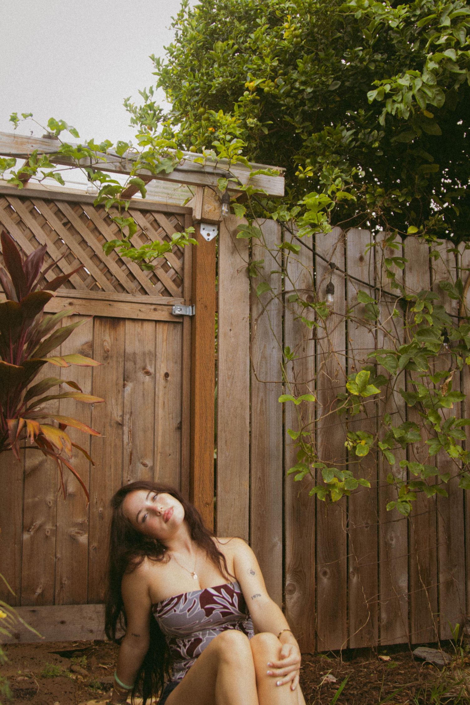 Young woman in a floral dress leaning against a wooden fence surrounded by green vines and tropical plants.