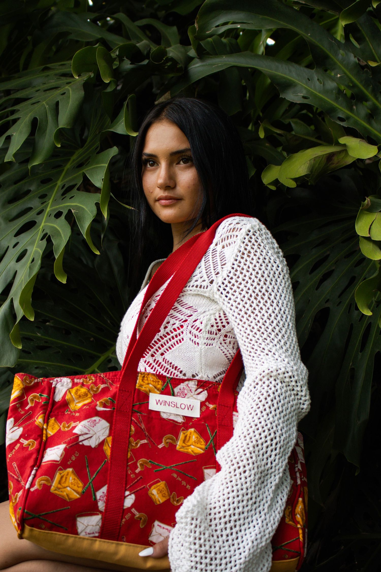 Dark-haired woman posing with a red Winslow patterned tote bag against lush tropical monstera leaves.