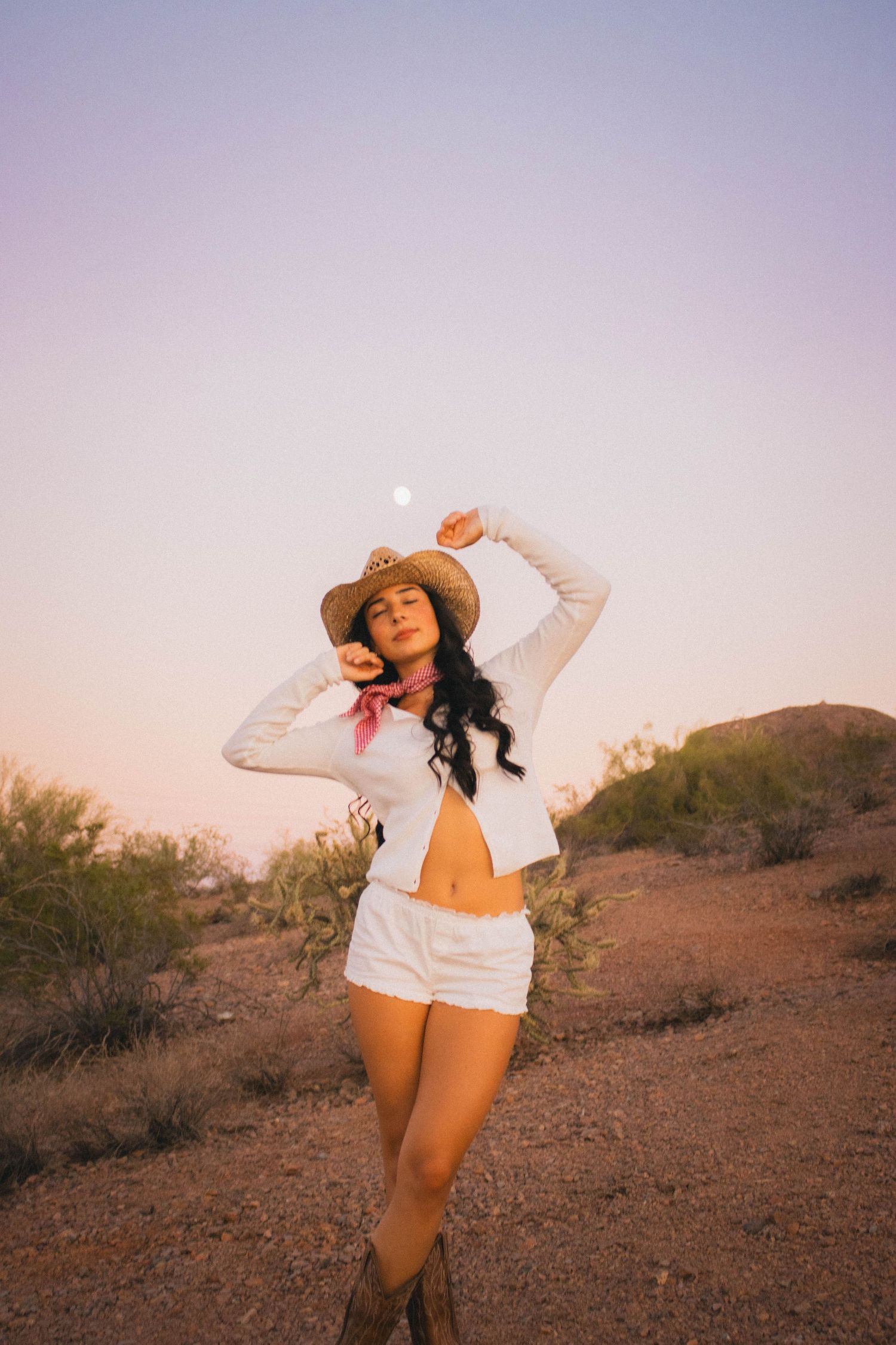 Woman in white outfit and cowboy hat poses in desert landscape during golden hour.