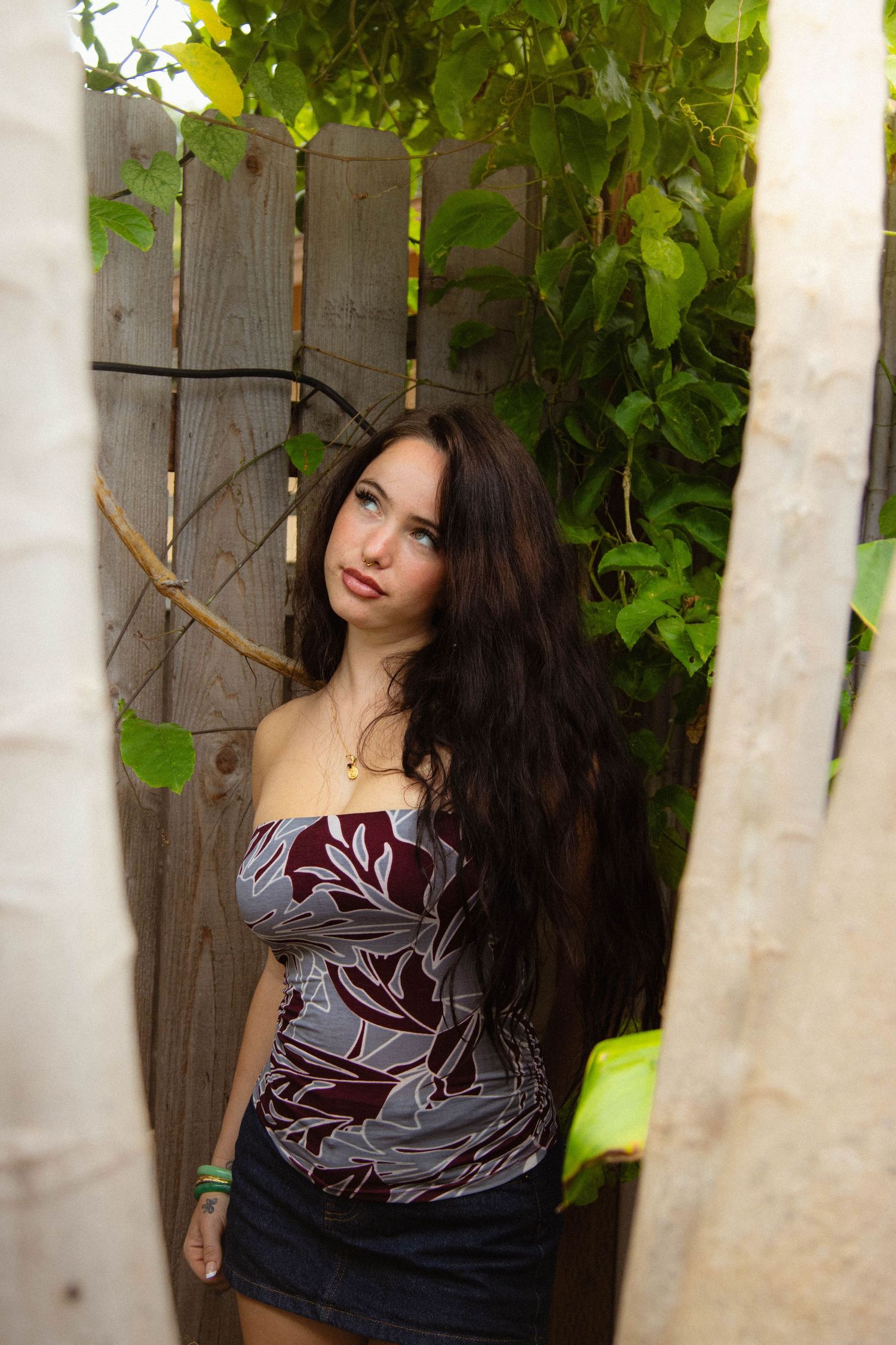 Young woman with long dark hair in a floral tube top posing between wooden posts surrounded by green vines.