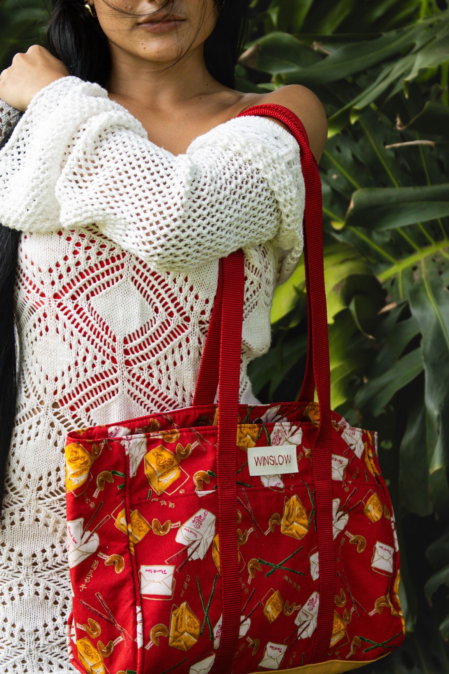 Model carrying a red Winslow branded tote bag with food print pattern, wearing a white knit sweater outdoors.