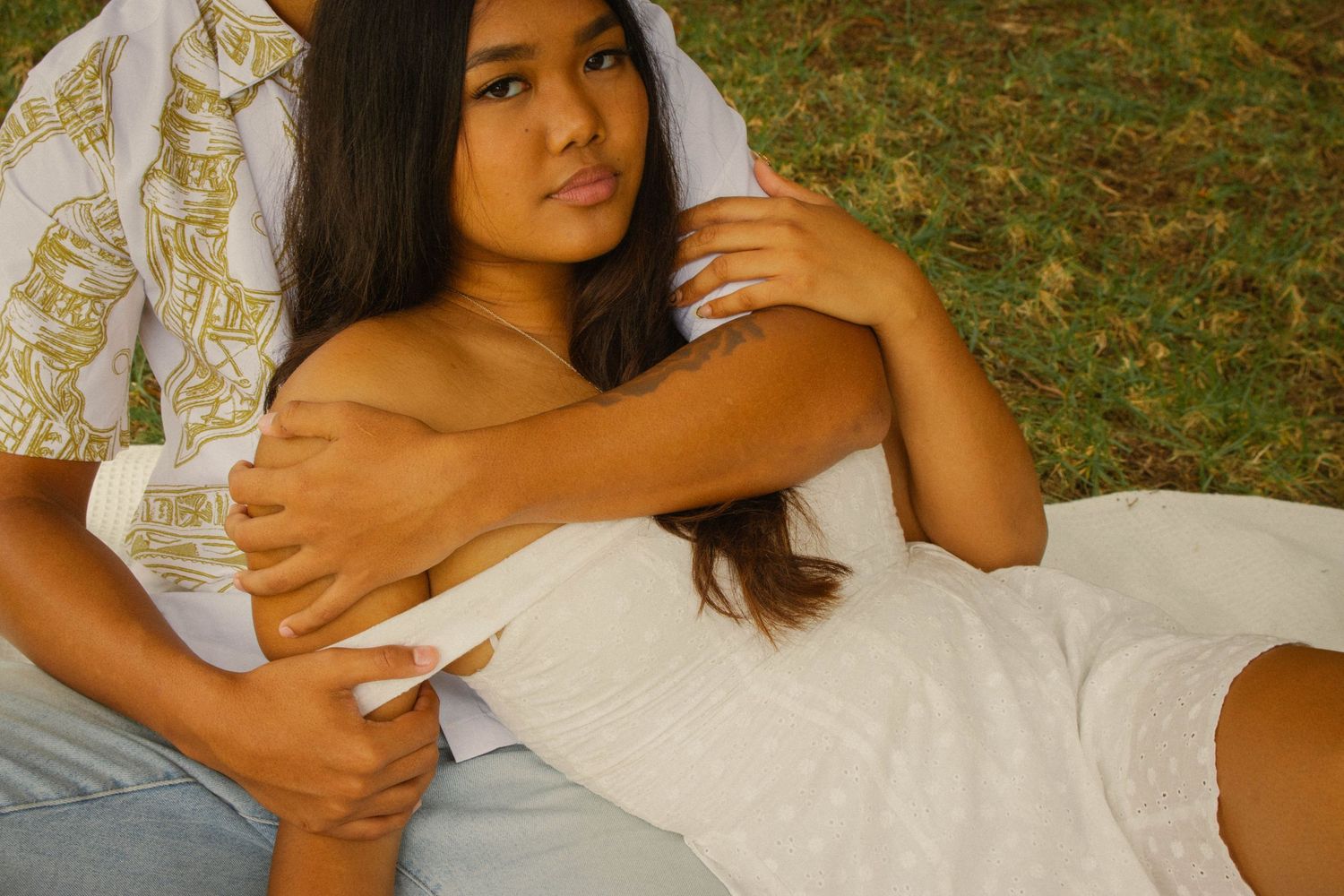 Young woman in a white dress lying on grass, leaning against a man in a patterned shirt outdoors.
