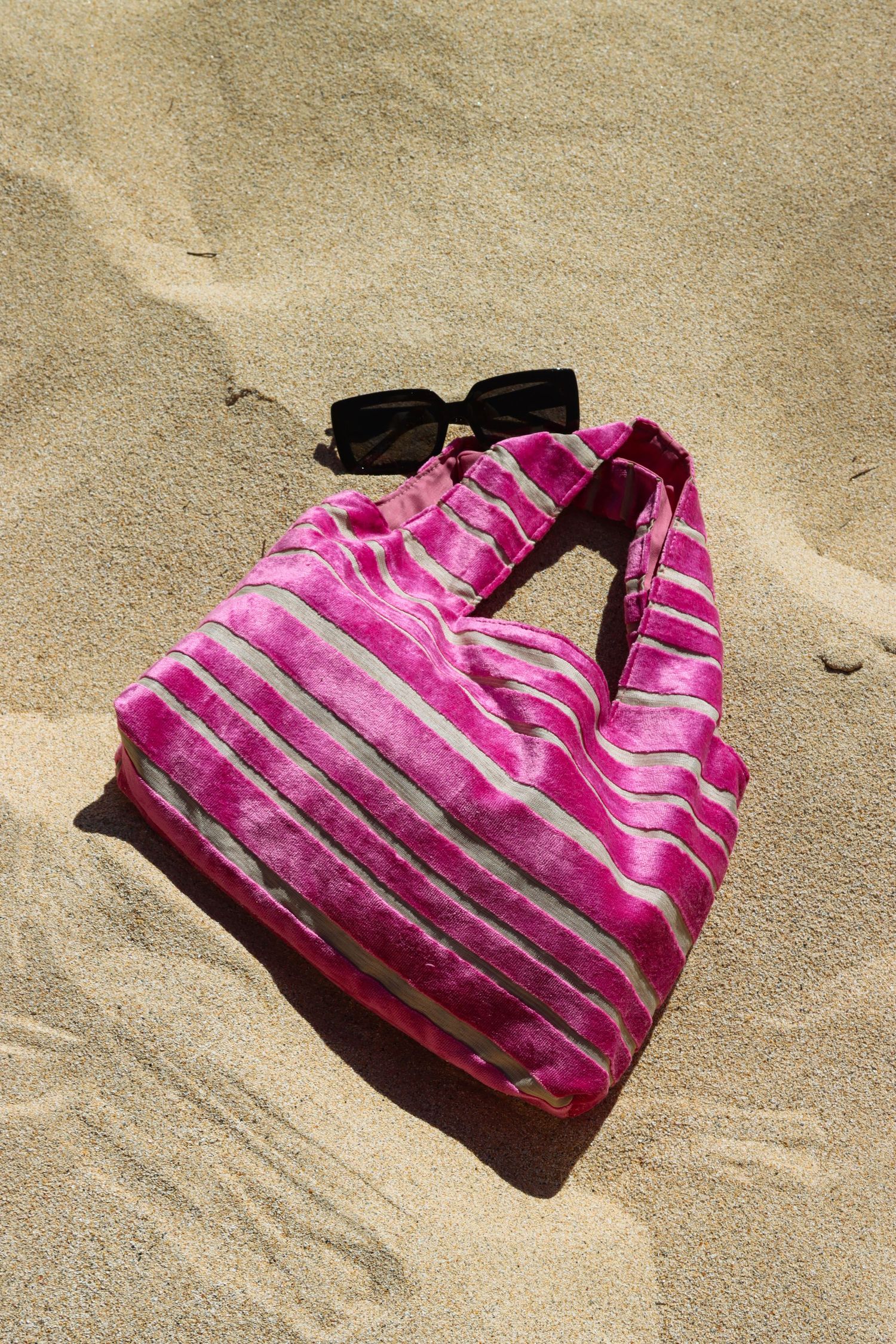 Pink striped beach tote bag with black sunglasses resting on sandy beach in bright sunlight.