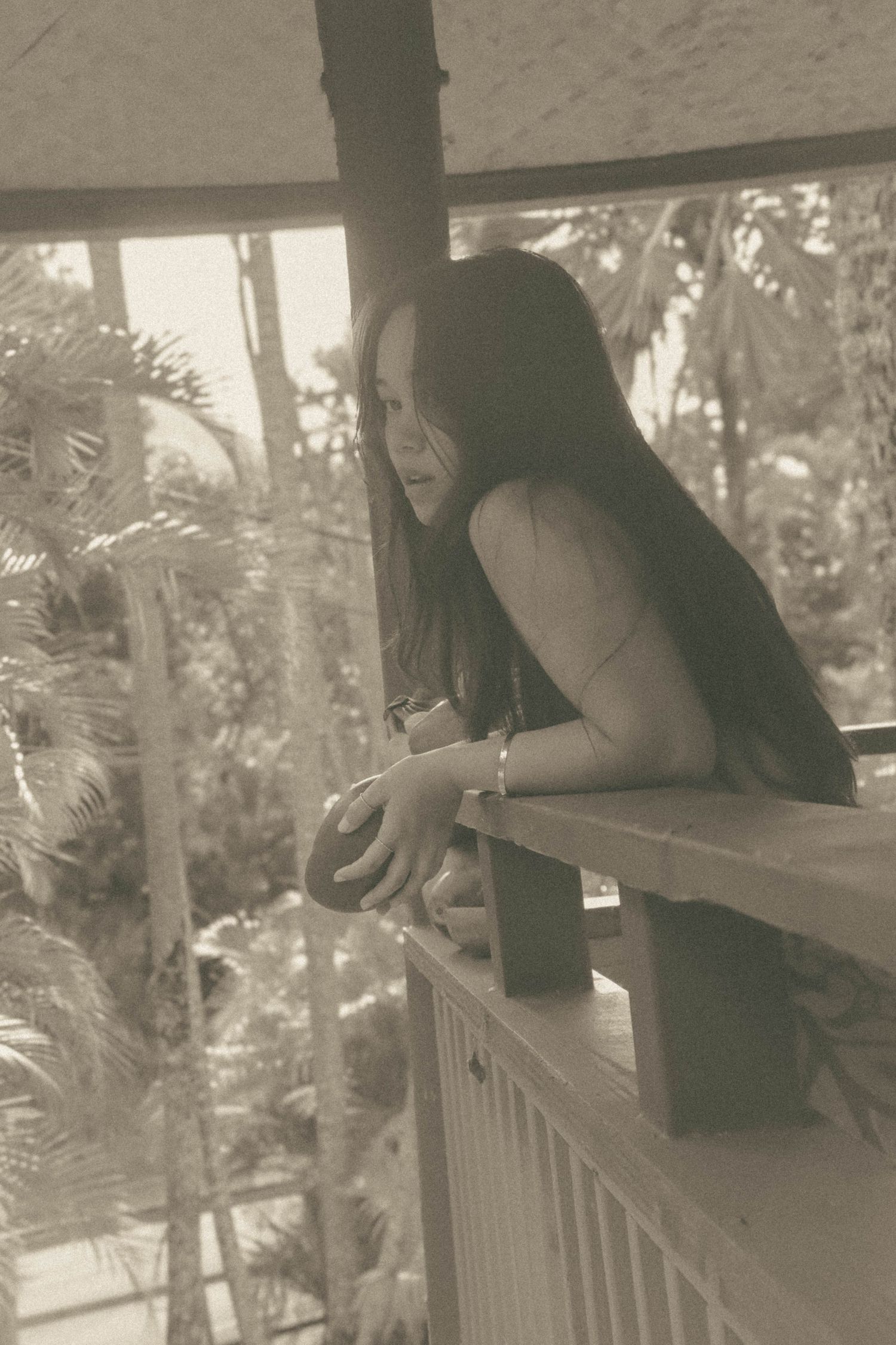Sepia toned photo of long-haired woman leaning on wooden balcony railing overlooking tropical palms.