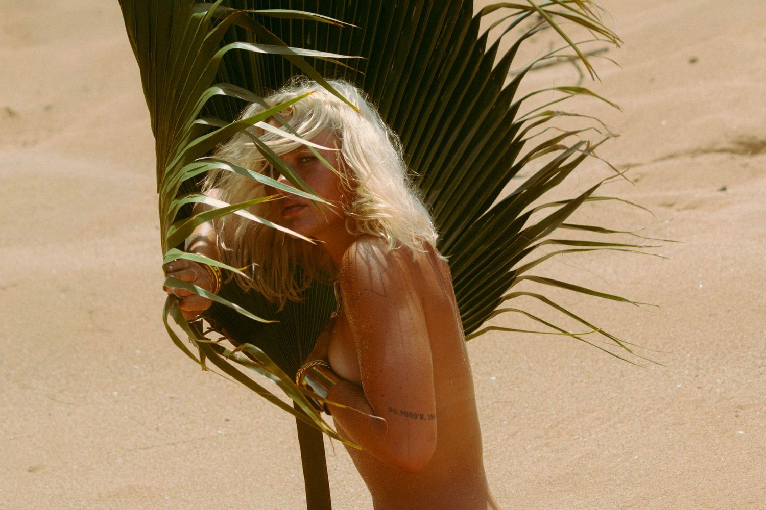 Blonde woman partially obscured by large green tropical palm leaf on sunny sandy beach.