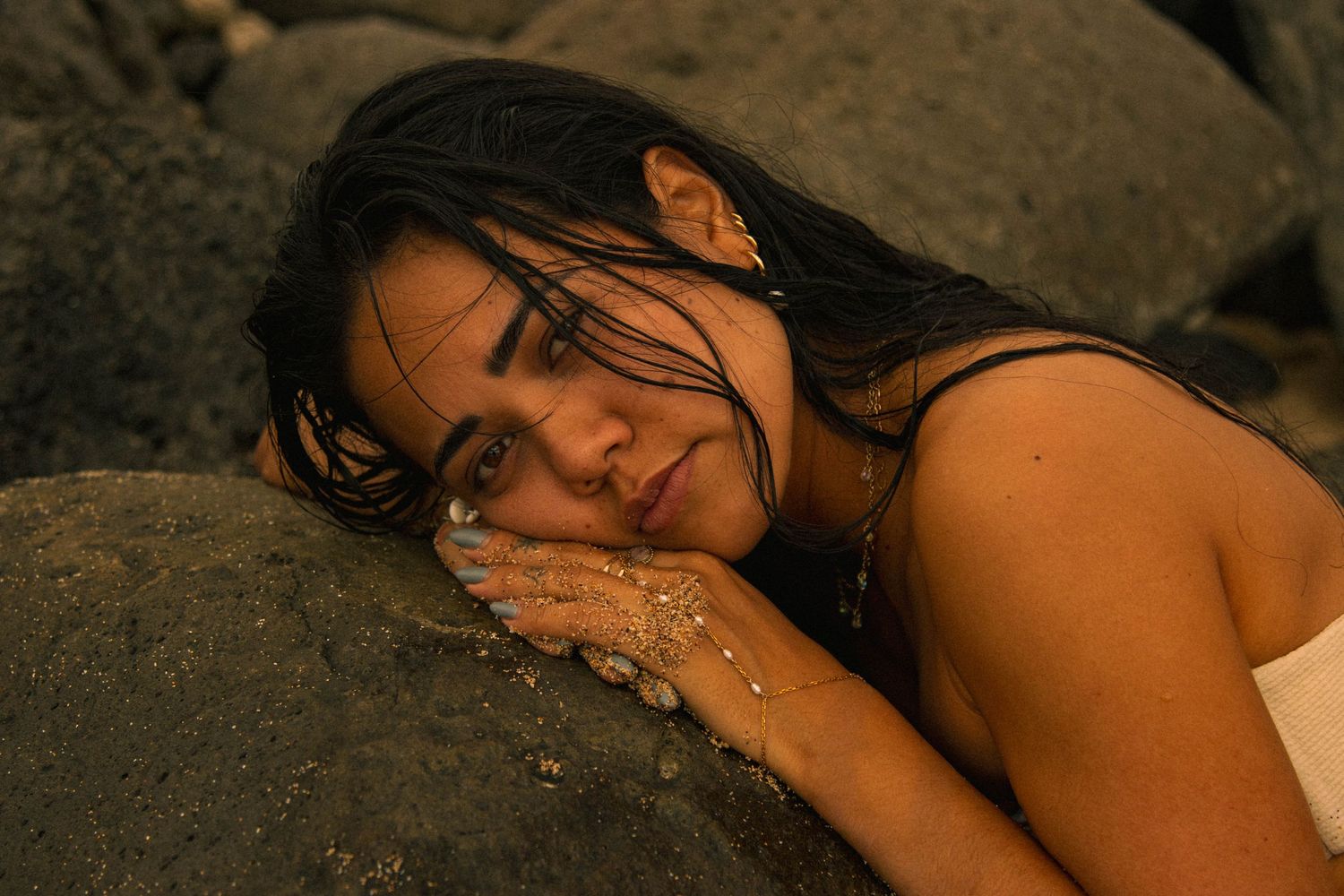 Dark-haired woman resting her face on a rock at the beach in warm golden light.