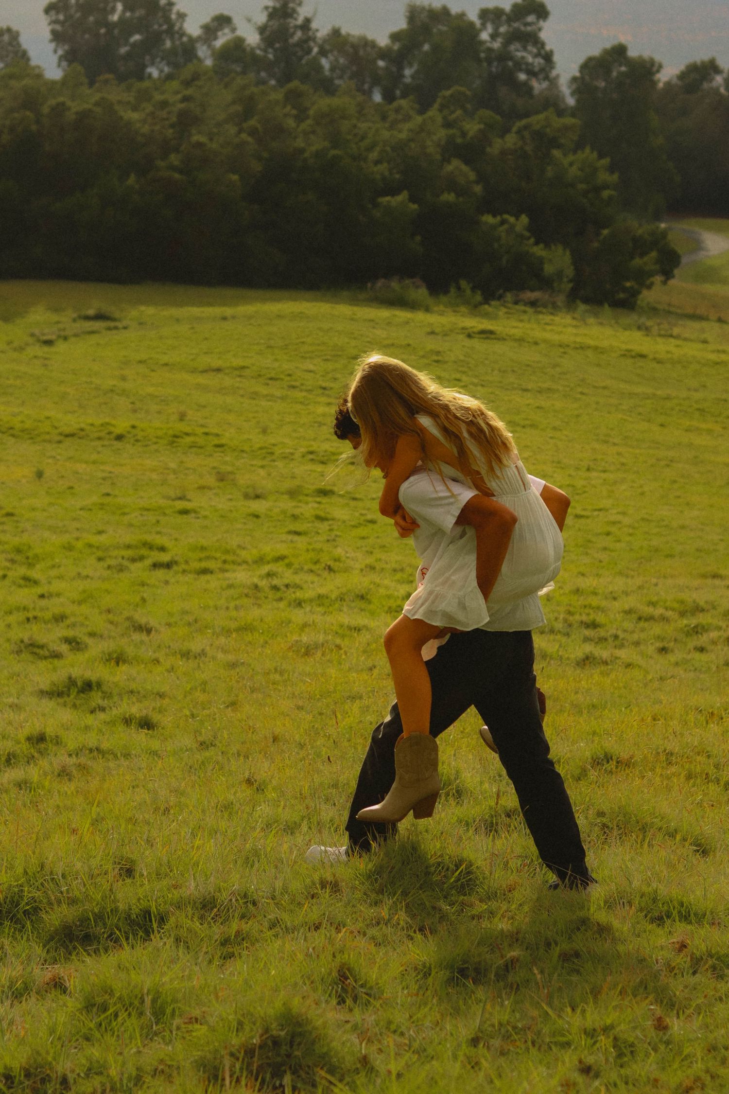 Man giving woman a piggyback ride across a sunny green field with trees in background.