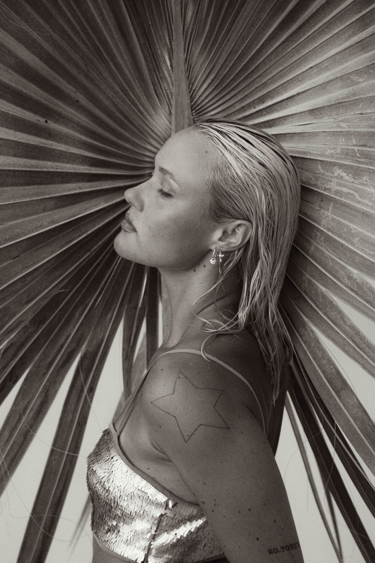 Black and white profile portrait of blonde woman leaning against large dried tropical palm leaf.